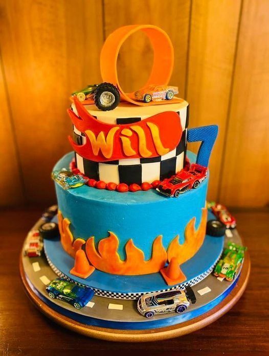 Tiered blue-and-red race car birthday cake with toy cars, flames, and a large orange “7” topper