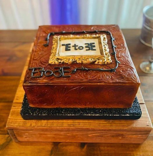 Chocolate cake decorated like a square leather trunk on a wooden board, with “E to 3E” icing on top.