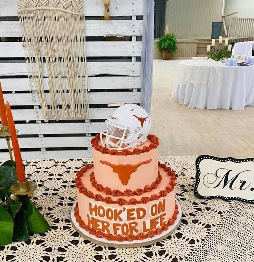 Orange and white football-themed cake with a helmet topper and “HOOK’D ON HER FOR LIFE” text at a wedding display