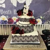 Three-tier white and black wedding cake with red roses and a black topper on a decorated table.