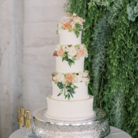Three-tier white wedding cake with peach flowers on a silver stand beside gold champagne flutes