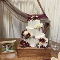 White wedding cake with burgundy flowers on a wooden crate, set against draped fabric backdrop