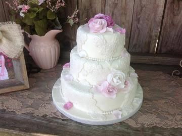 Three-tier white wedding cake with pink roses on a round white stand, displayed on a rustic table