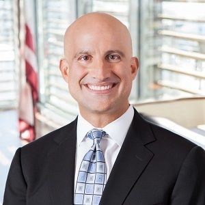 Bald man in a suit smiling, standing in front of windows with an American flag.