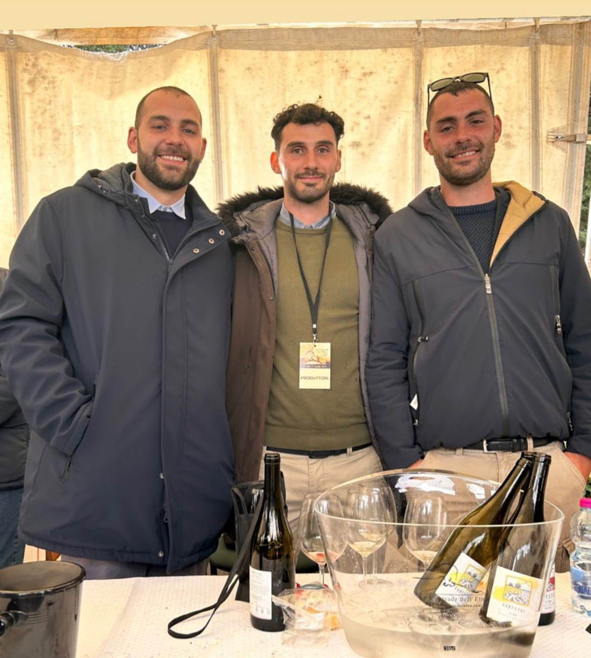 Tre uomini in piedi dietro un tavolo con bottiglie di vino e bicchieri. Uno indossa una giacca blu, uno una camicia verde, uno una felpa grigia con cappuccio.