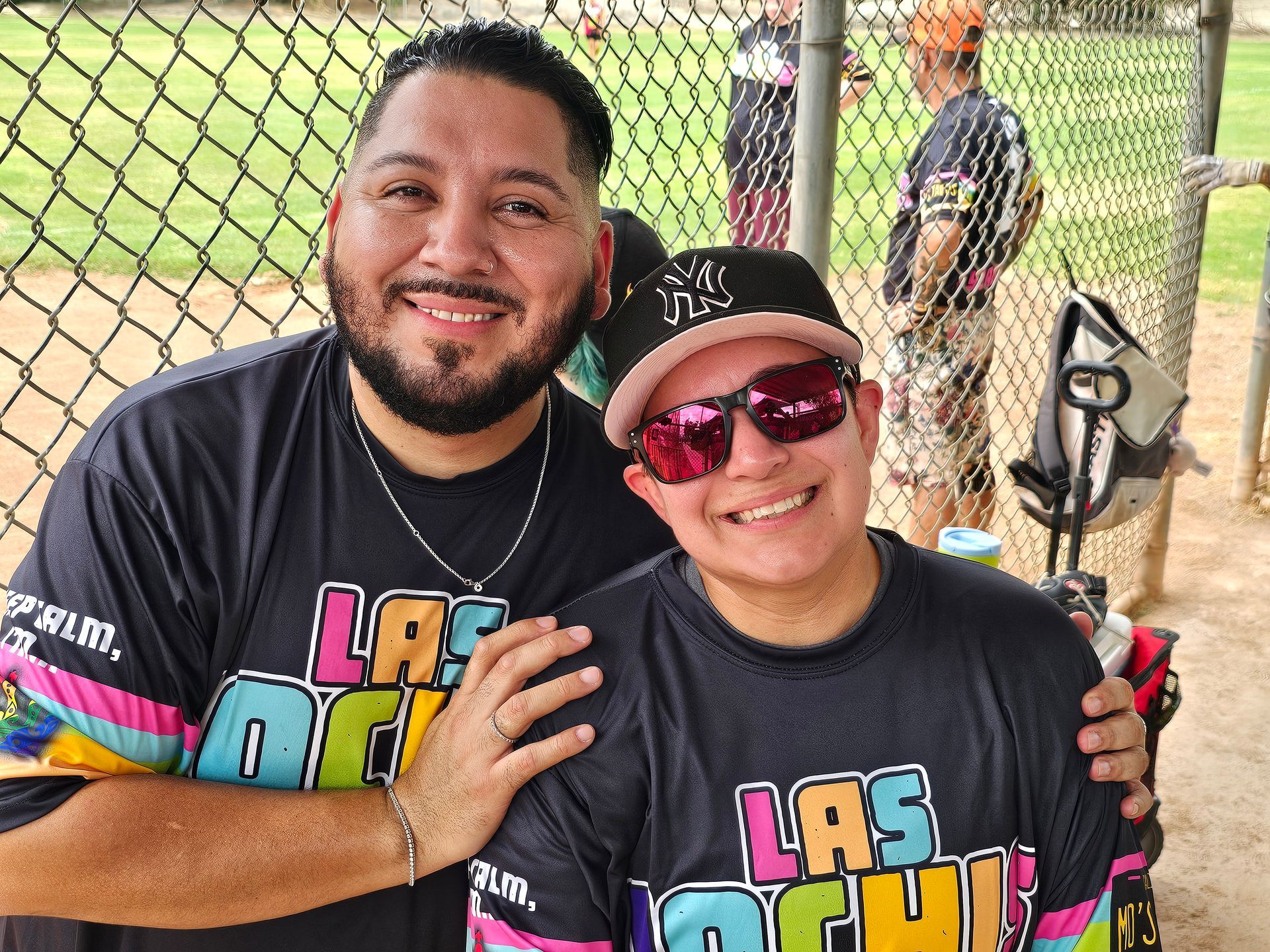 Two smiling people in matching black shirts pose at a baseball field fence.
