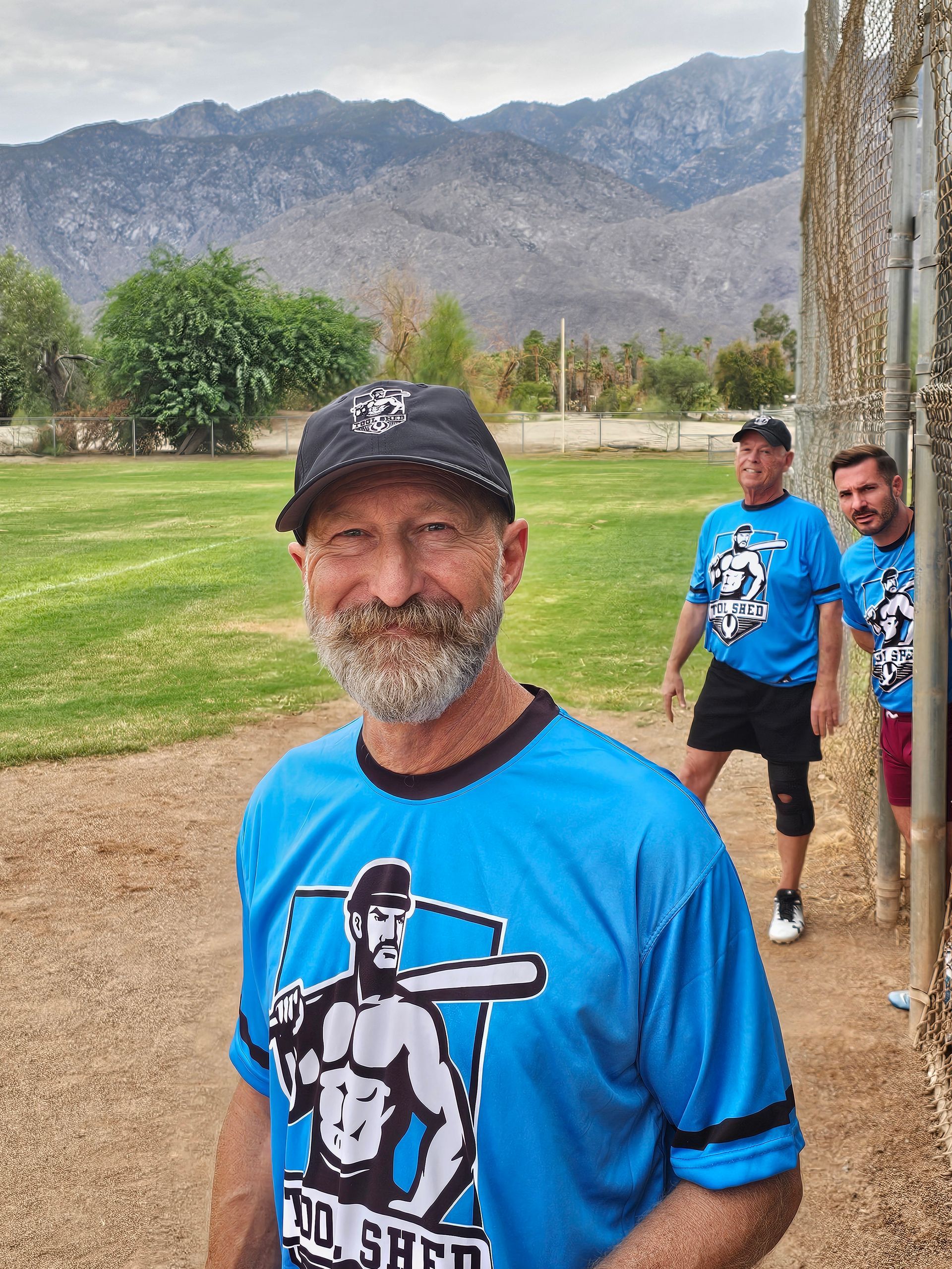 Smiling man in blue sports jersey and black cap stands on a dirt field with mountains behind him.