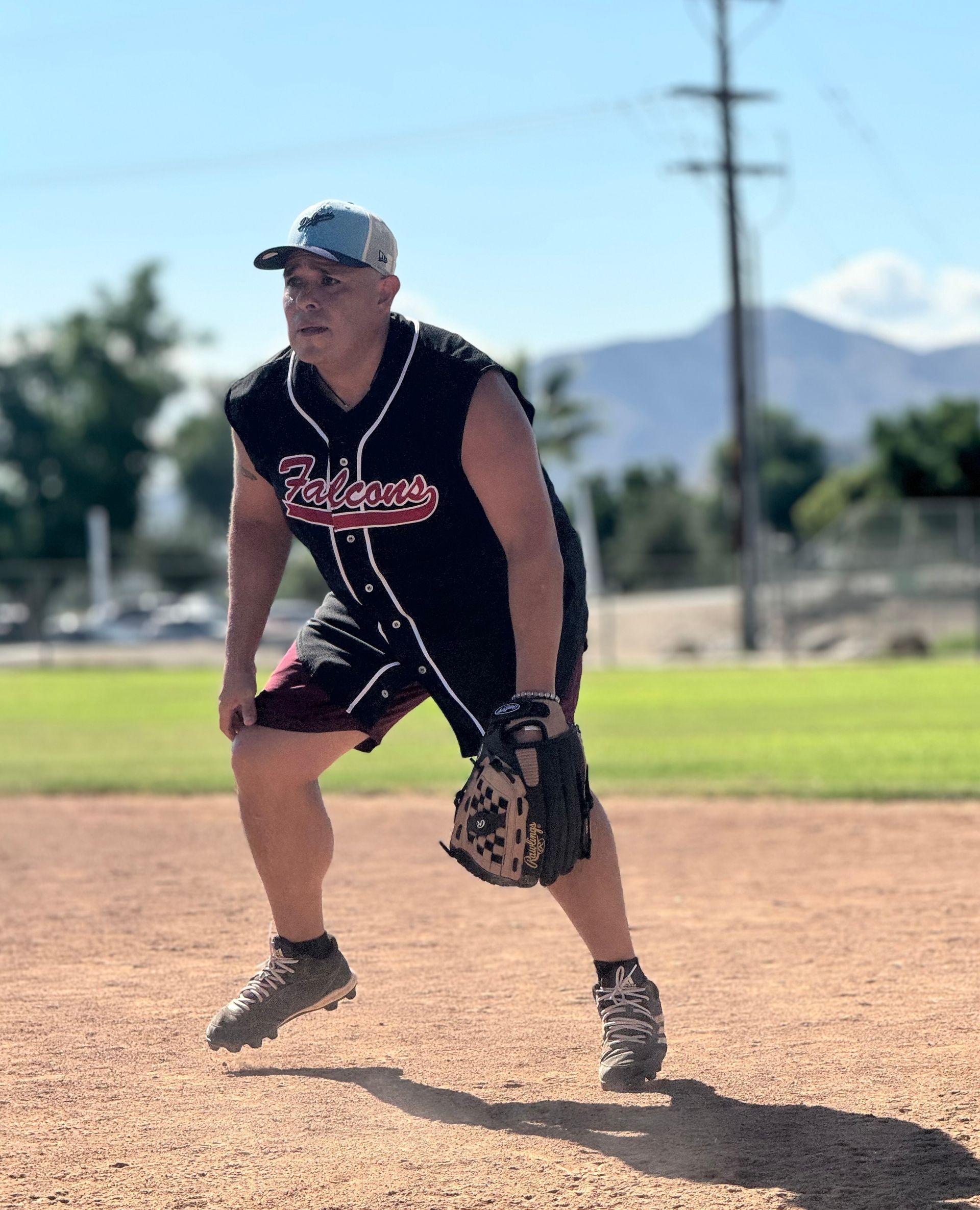 Softball player in black uniform crouches on a dirt field, ready with glove under a sunny sky.