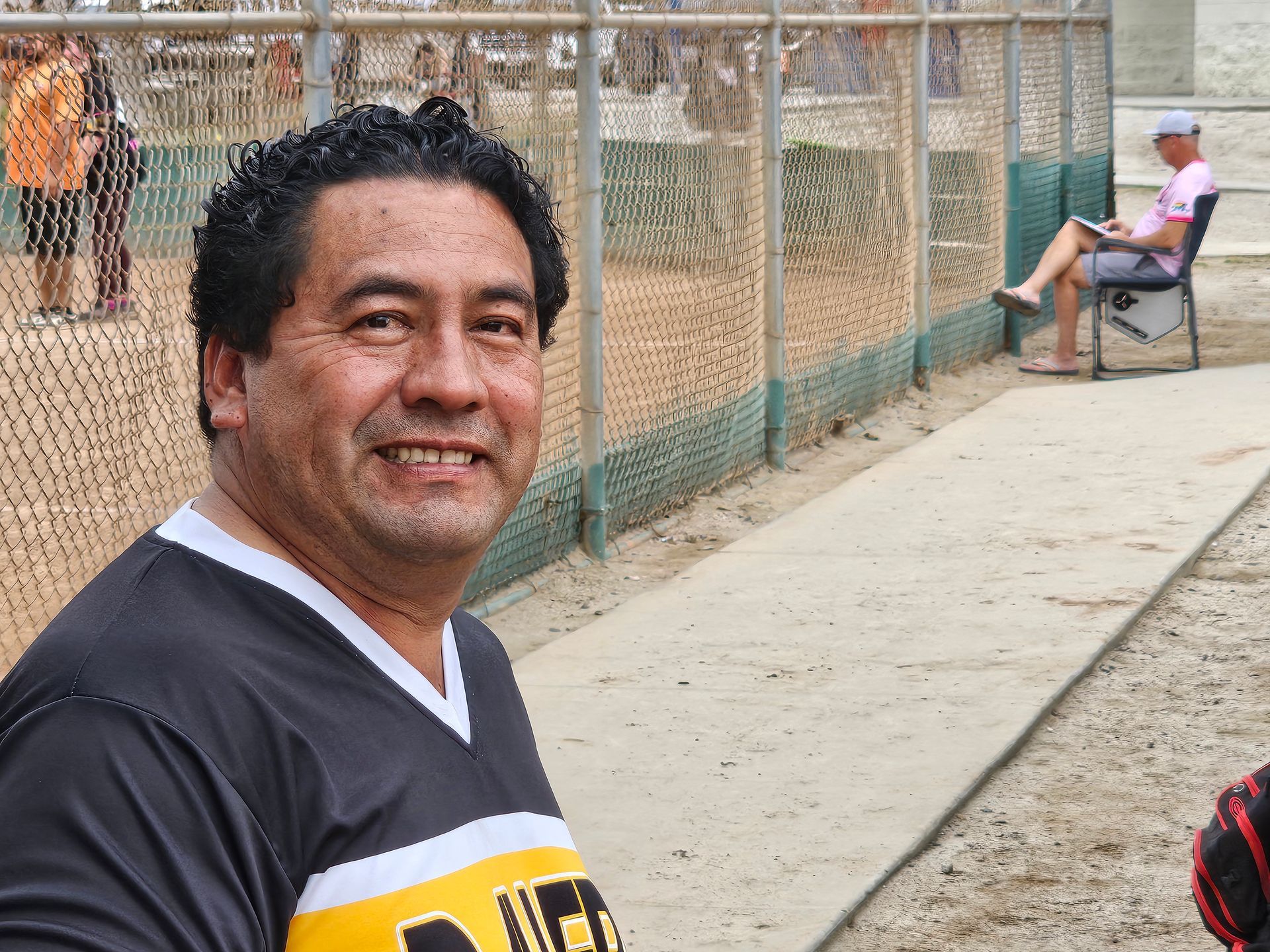 Man smiling beside a chain-link fence at an outdoor court, with a seated person in the background.