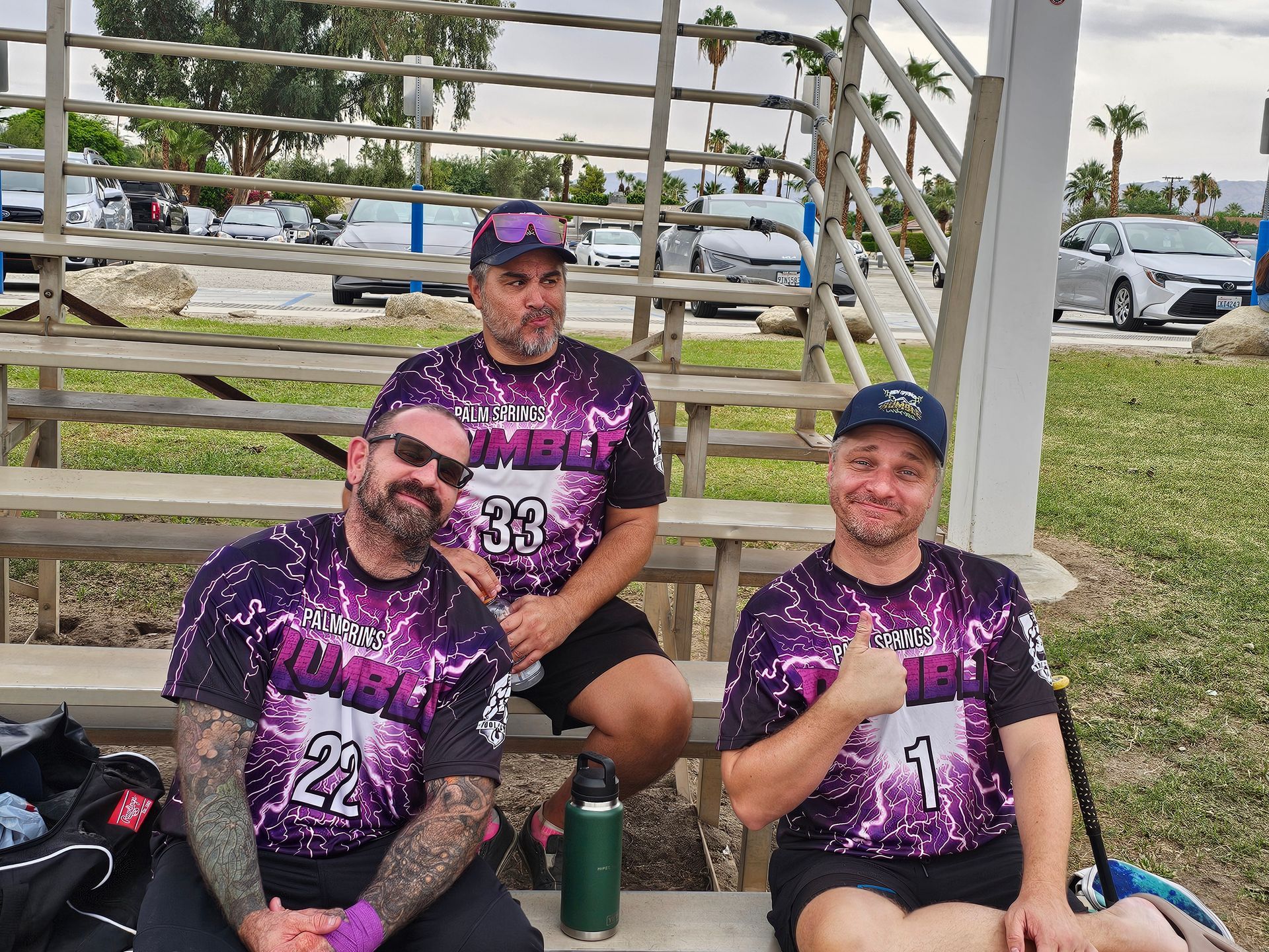 Three people in purple sports jerseys sit on a bench at an outdoor field, smiling and posing.