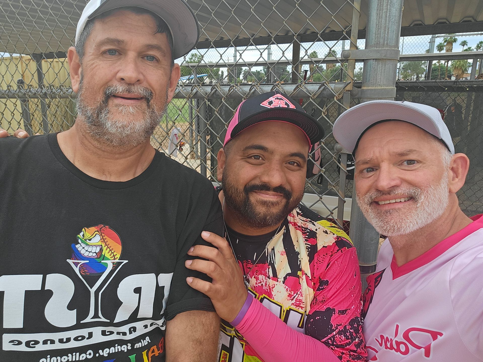 Three smiling people posing together outdoors by a fence, two in pink shirts and one in a black shirt.