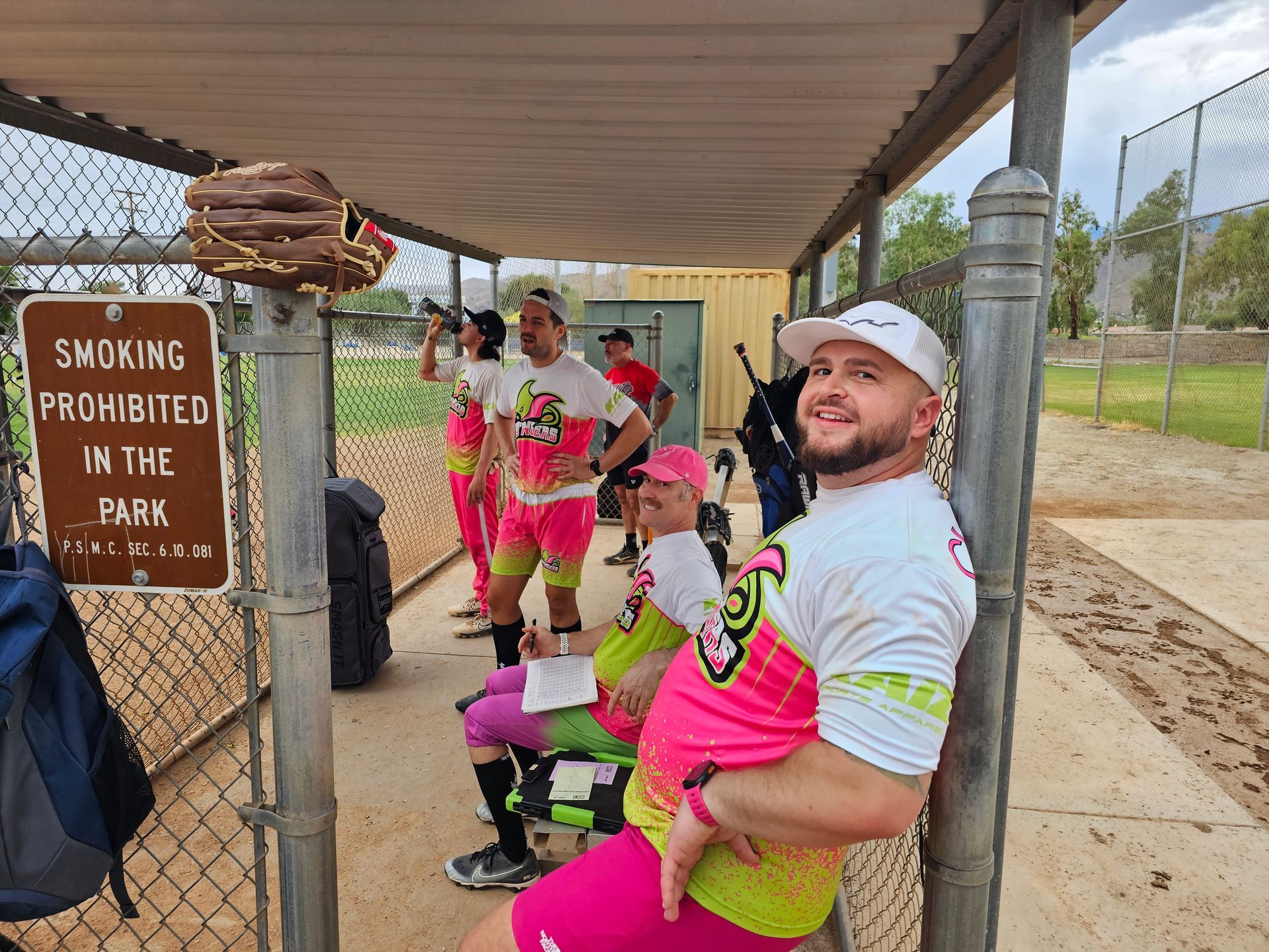 People in neon pink shirts sitting and standing under a shelter by a sign reading “Smoking prohibited in the park.”