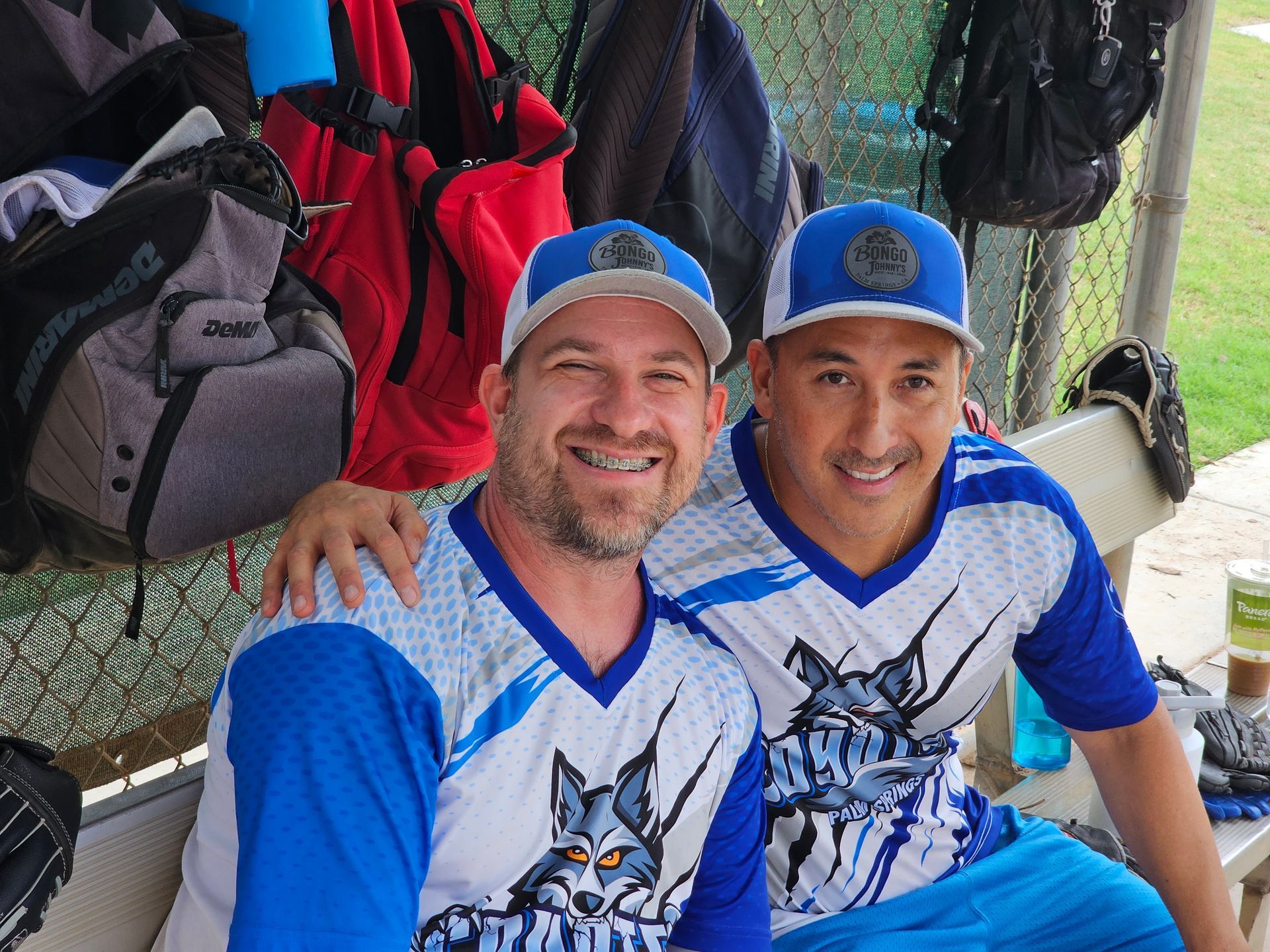 Two smiling teammates in blue baseball jerseys and caps posing together on a bench