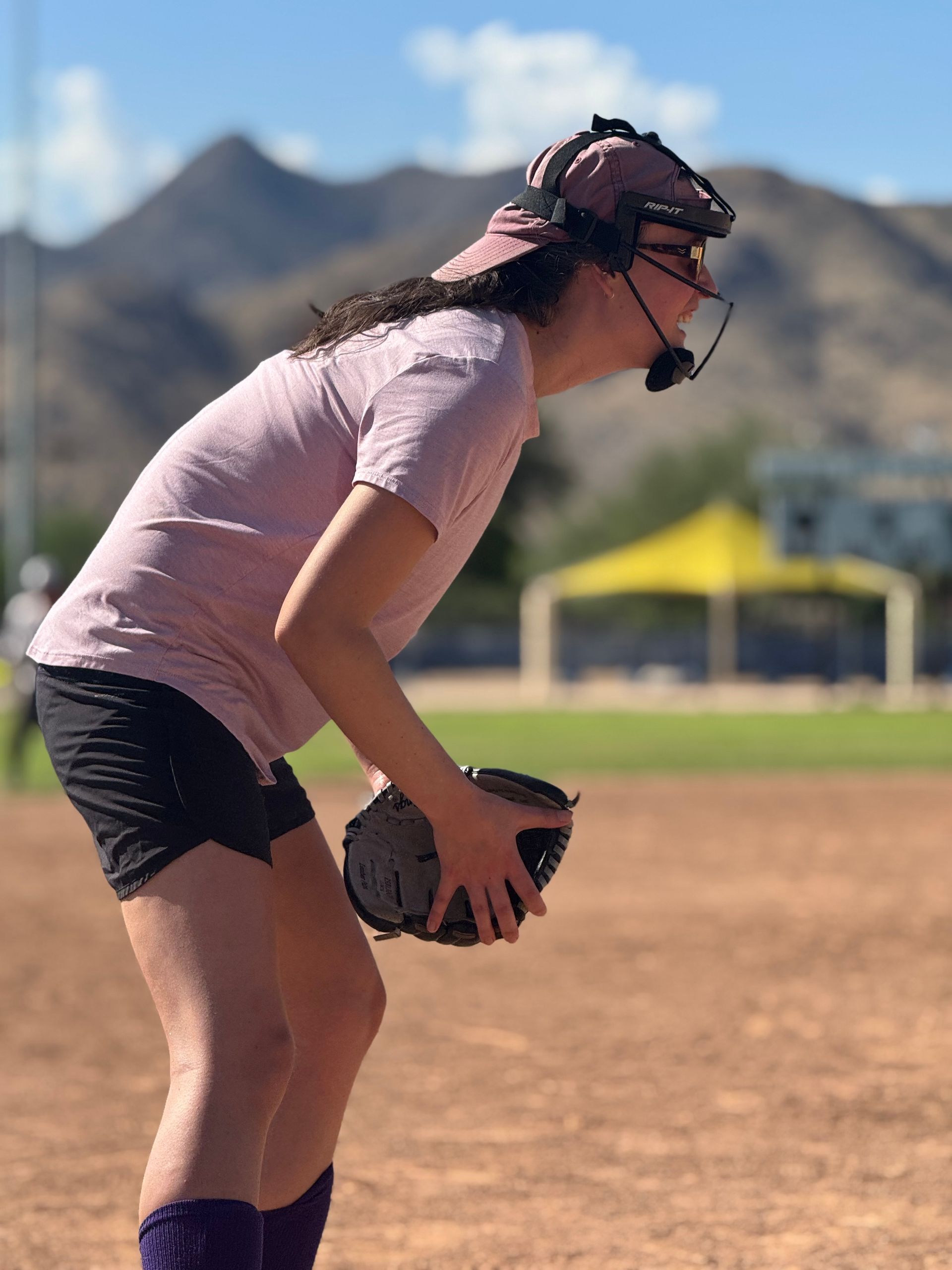 Softball player in pink shirt and black shorts bends forward holding a ball on a field with mountains behind.