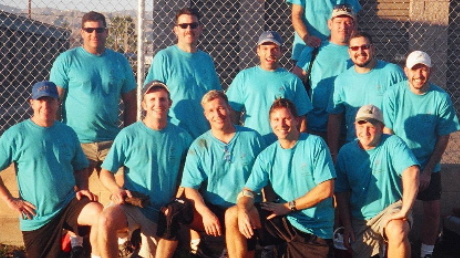 Group of baseball players in red jerseys posing outdoors on grass, with mountains in the background