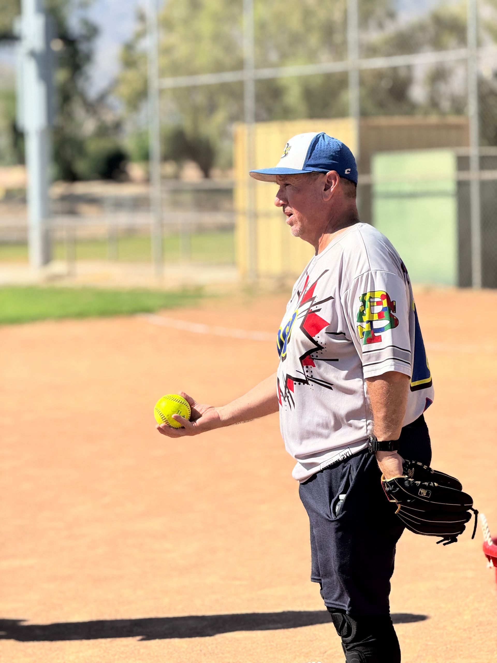 Softball player in gray jersey holding a yellow ball and glove on a sunny field