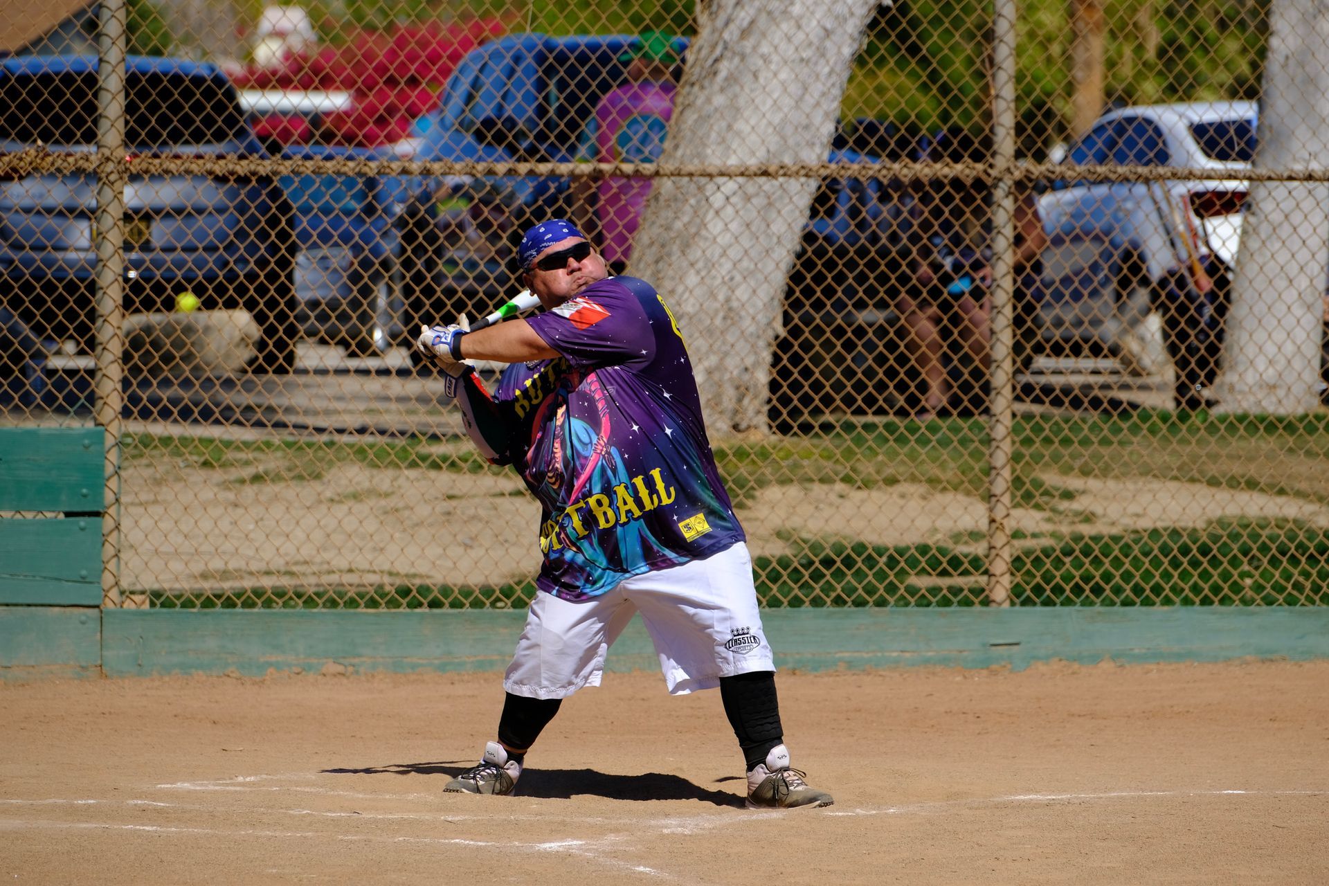 Baseball player in a colorful jersey swings a bat on a dirt field, with a chain-link fence and cars behind him.