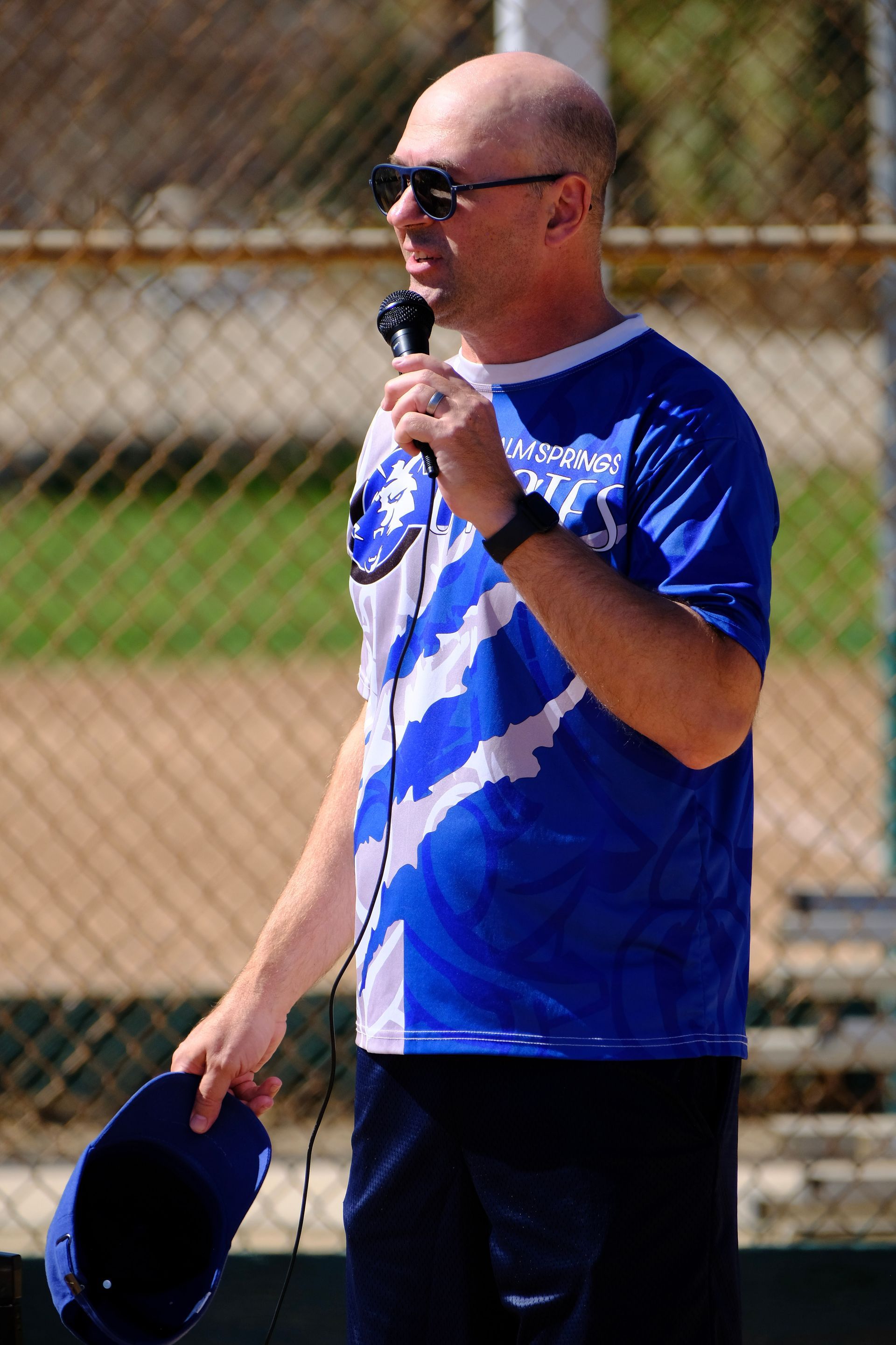 Man holding a microphone and cap on a sports field, wearing a blue jersey and sunglasses