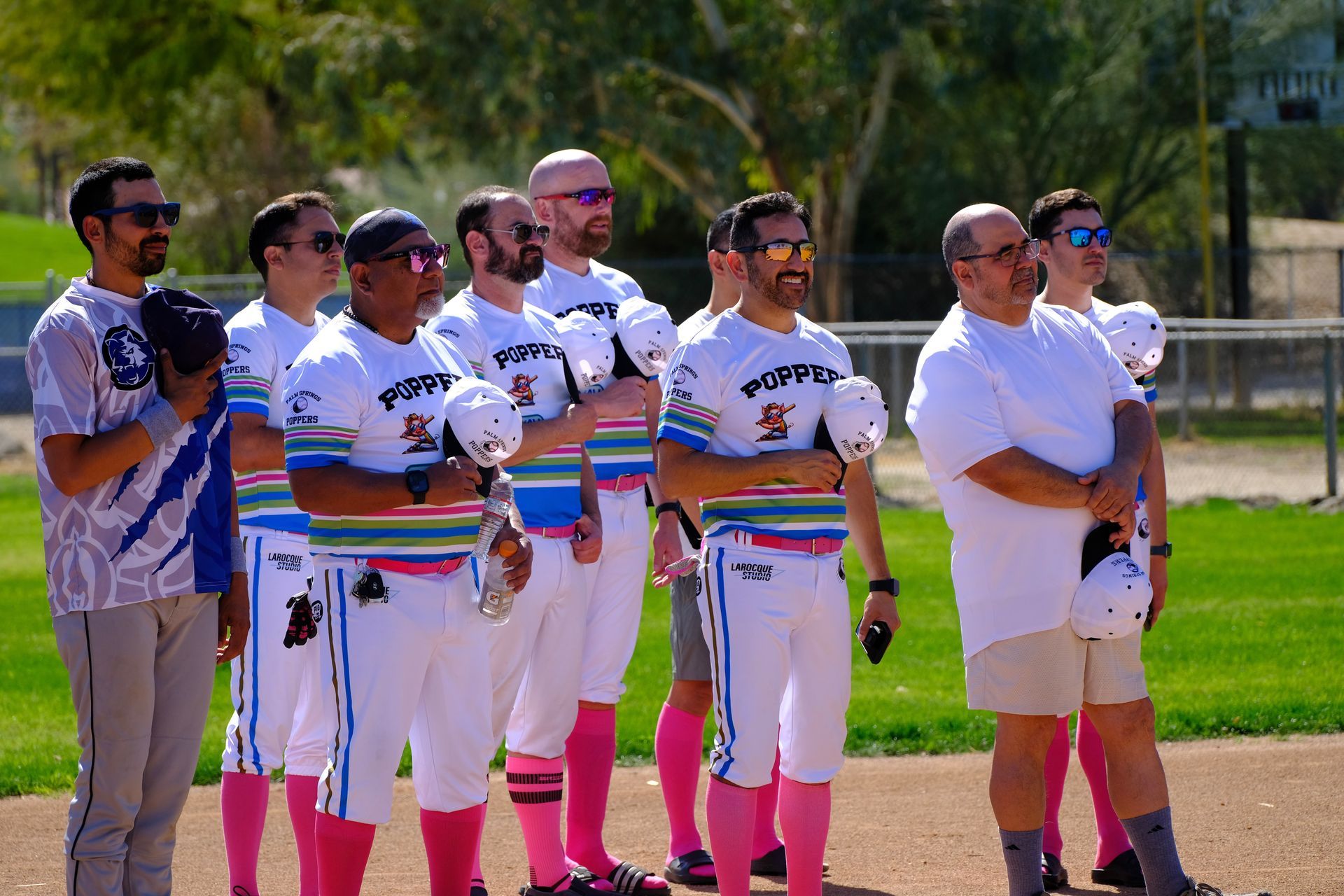 Softball team standing in a line on a field, wearing white uniforms and sunglasses, posing before a game