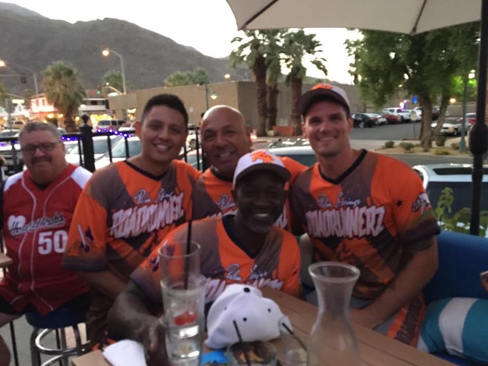 Five smiling friends in orange jerseys pose at an outdoor table under an umbrella at night.