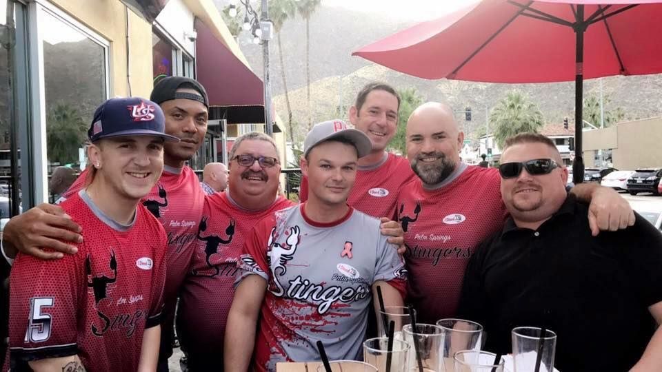 Group of people in red sports jerseys smiling at an outdoor table under red umbrellas