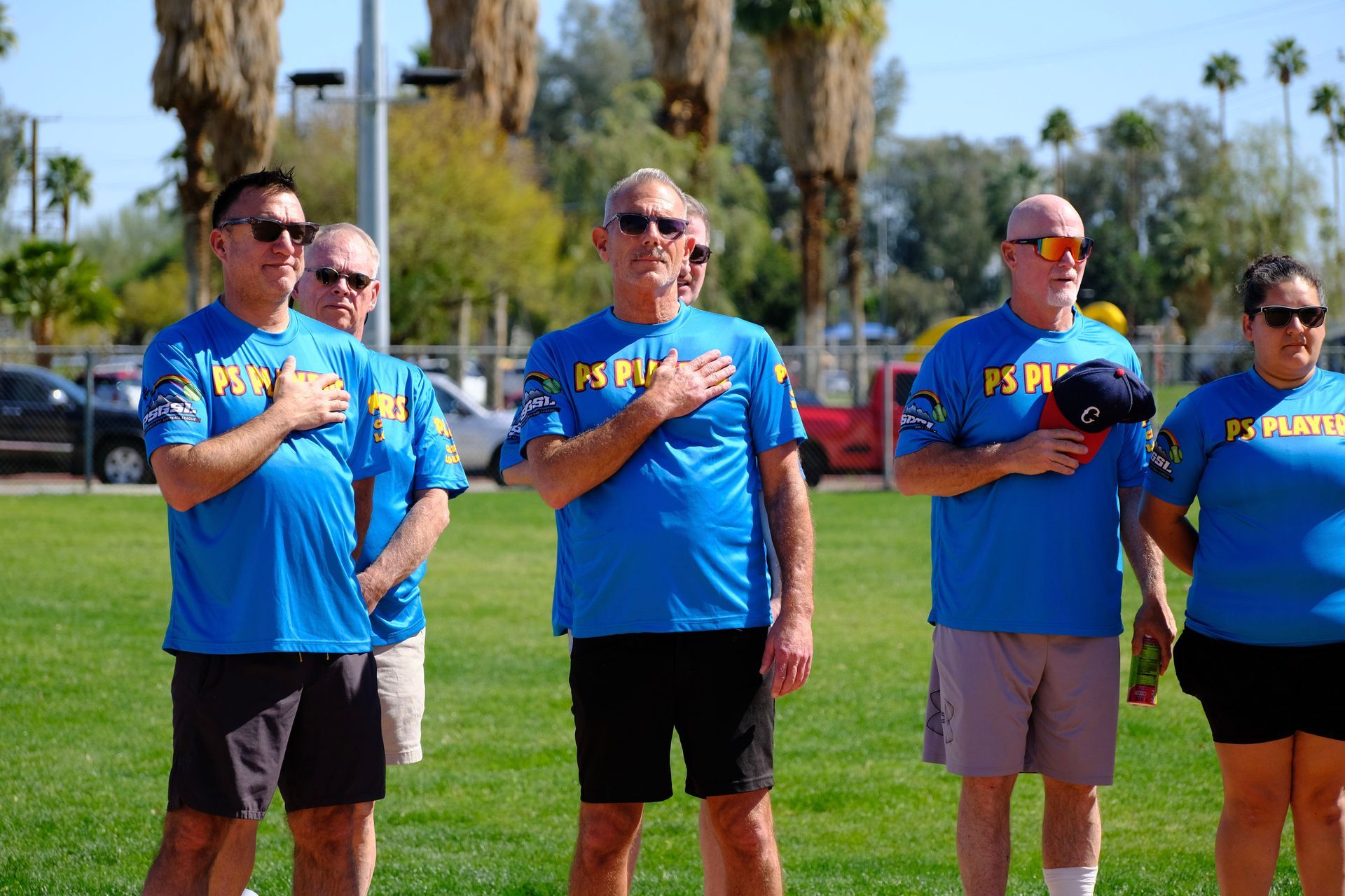 Four men in blue shirts stand outdoors with hands over their hearts during a ceremony.