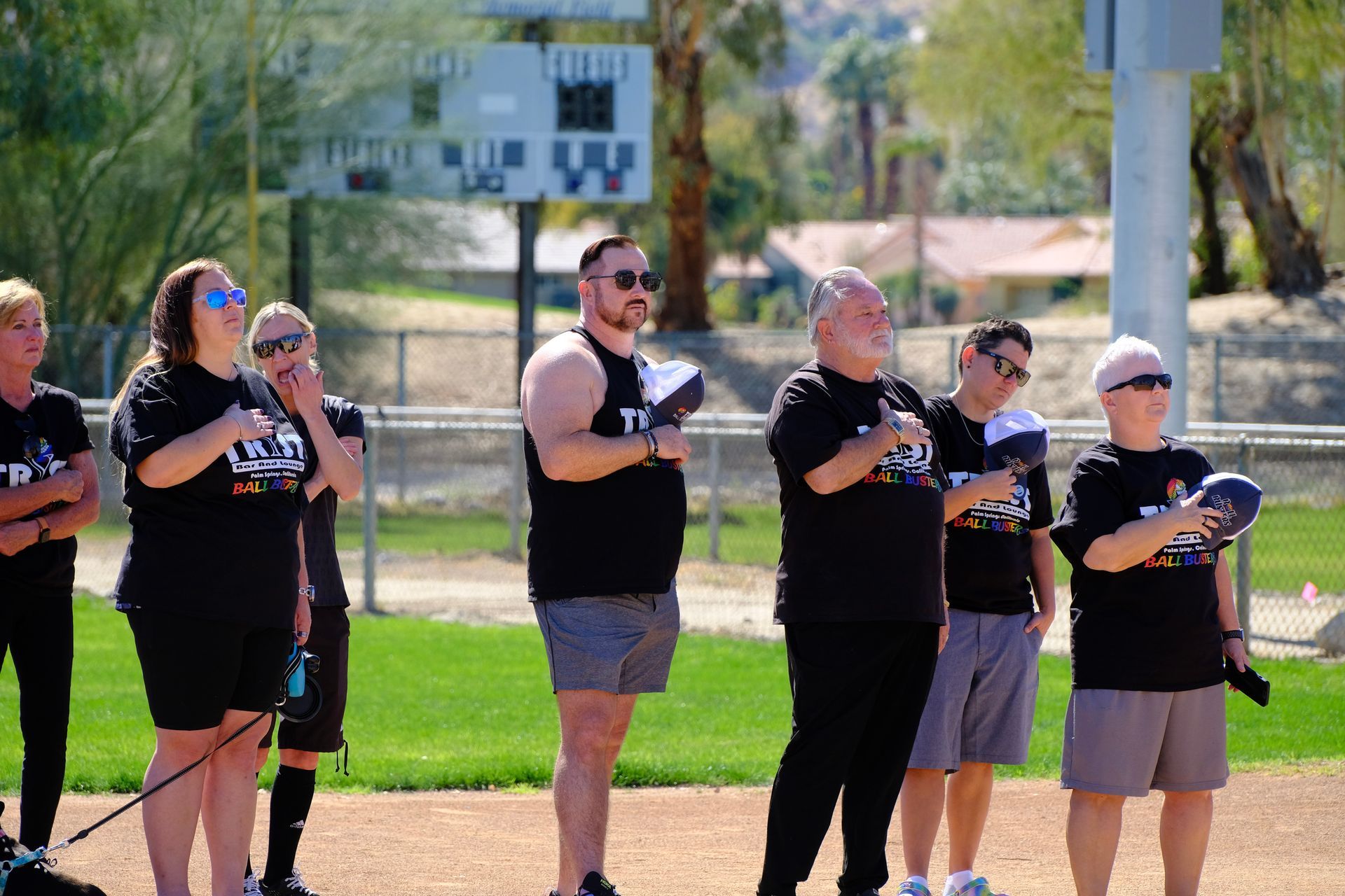 Six people in black shirts stand outdoors, clapping during a sunny event.