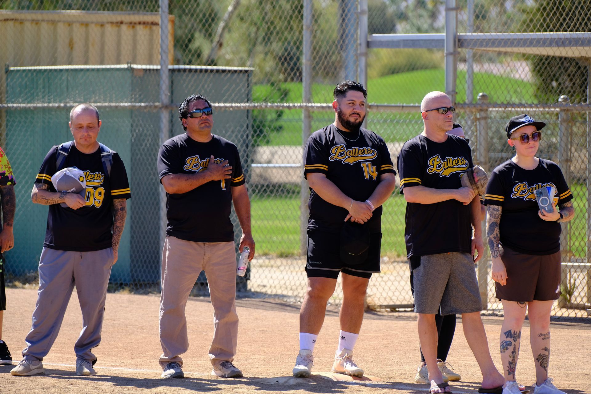 Five people in black shirts stand on a baseball field holding gloves, in front of a chain-link fence.