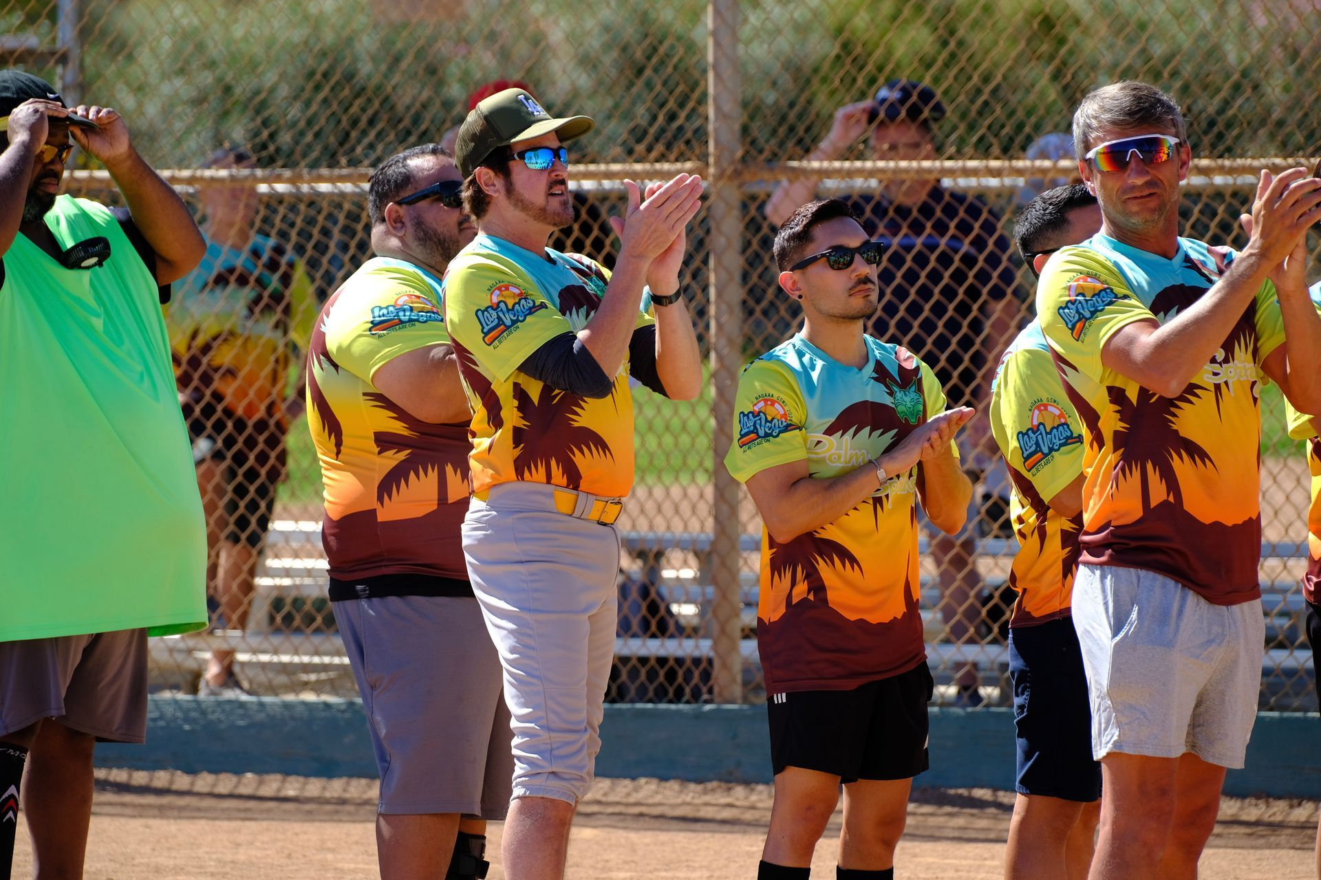 Fans in bright tie-dye shirts cheer and clap behind a fence at an outdoor event.