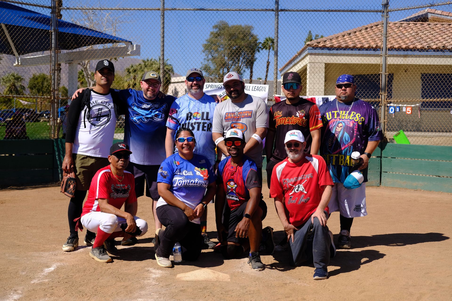 Softball team posing on a dirt field with bats and gloves in front of a chain-link fence.