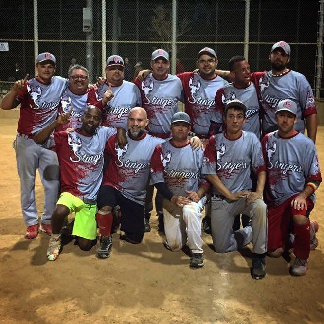 Softball team posing on a dirt field at night, wearing matching gray and red jerseys.