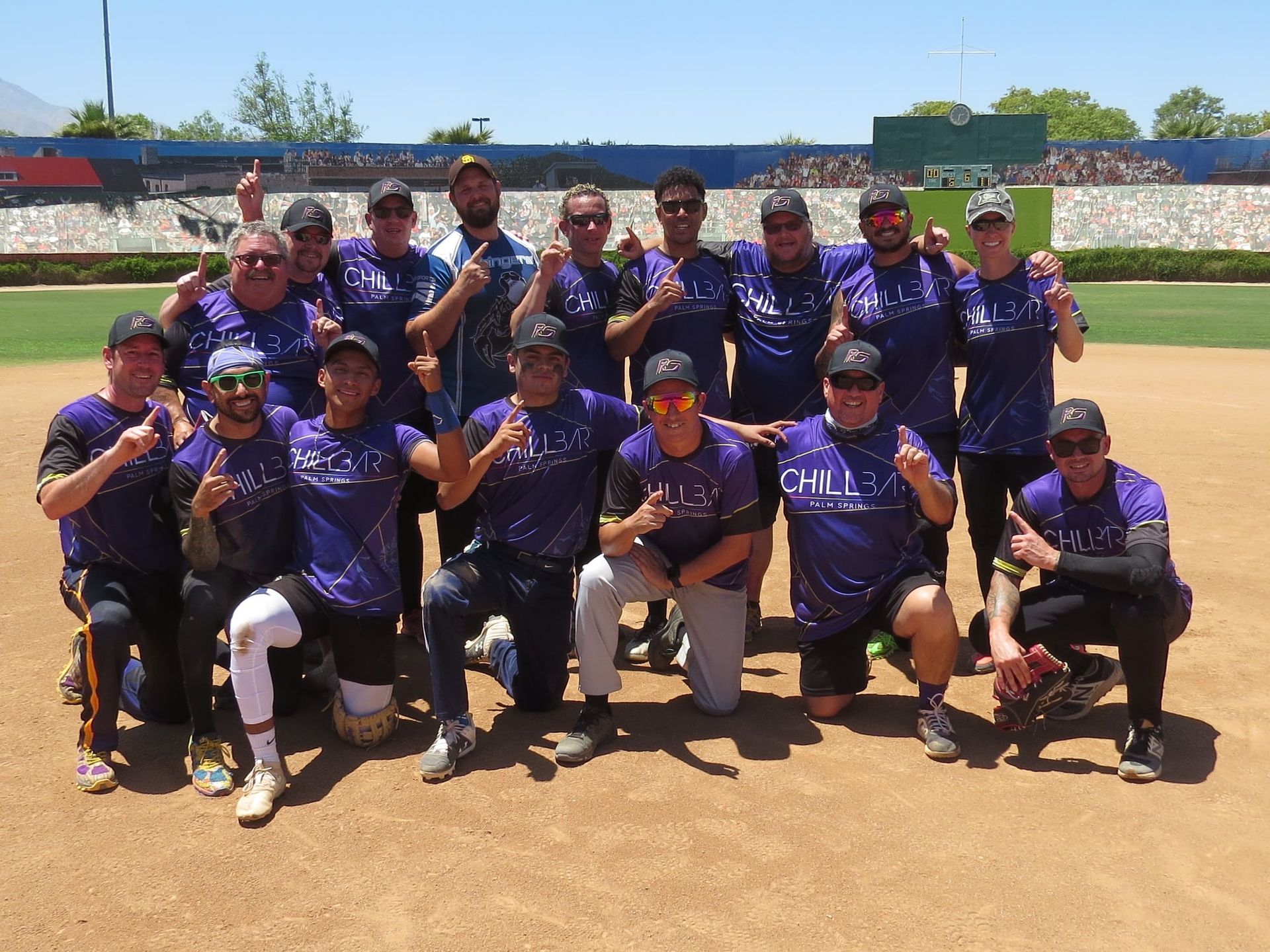 A softball team posing on a dirt field, wearing purple jerseys and caps, with gloves and bats.