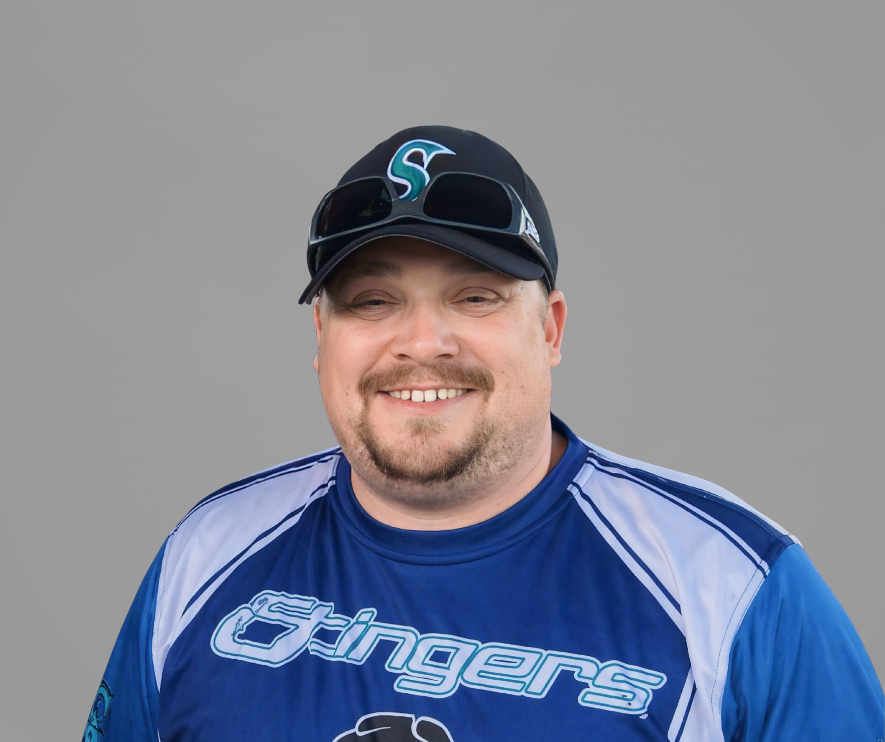 Smiling person in a blue baseball jersey and black cap against a gray background.