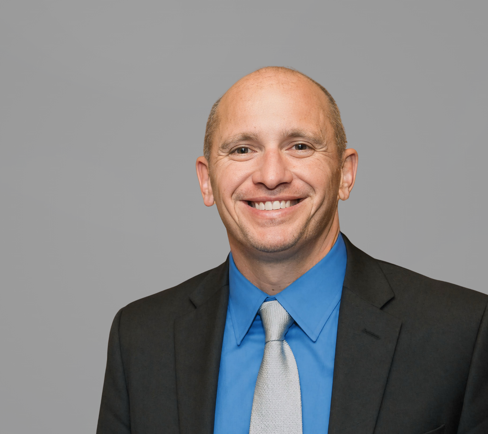 Smiling man in a black suit, blue shirt, and light tie against a gray background