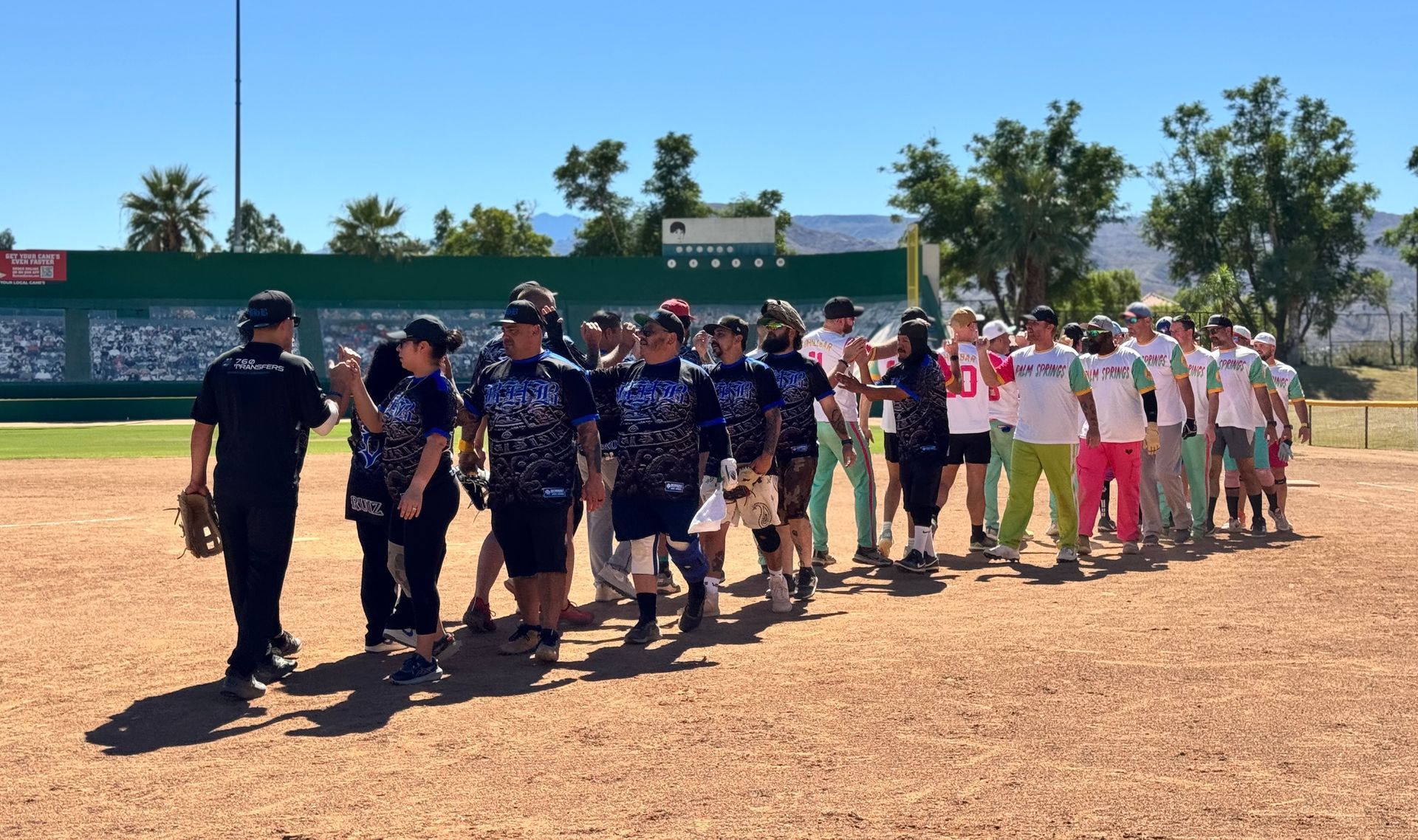 Two softball teams and coaches line up on a dirt field for a pregame handshake under a clear blue sky