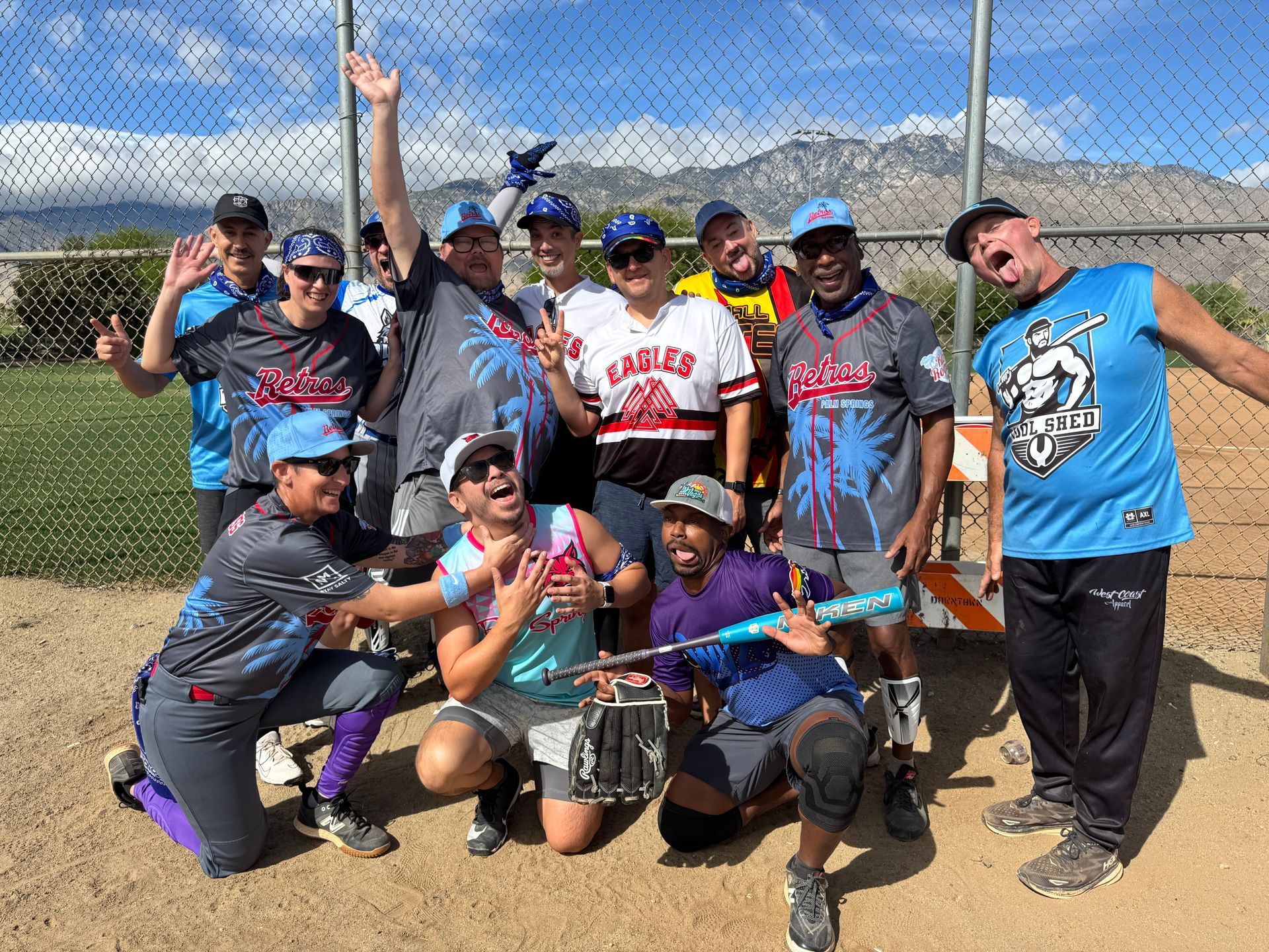 Group of softball players posing outdoors on a dirt field, smiling and celebrating with arms raised.