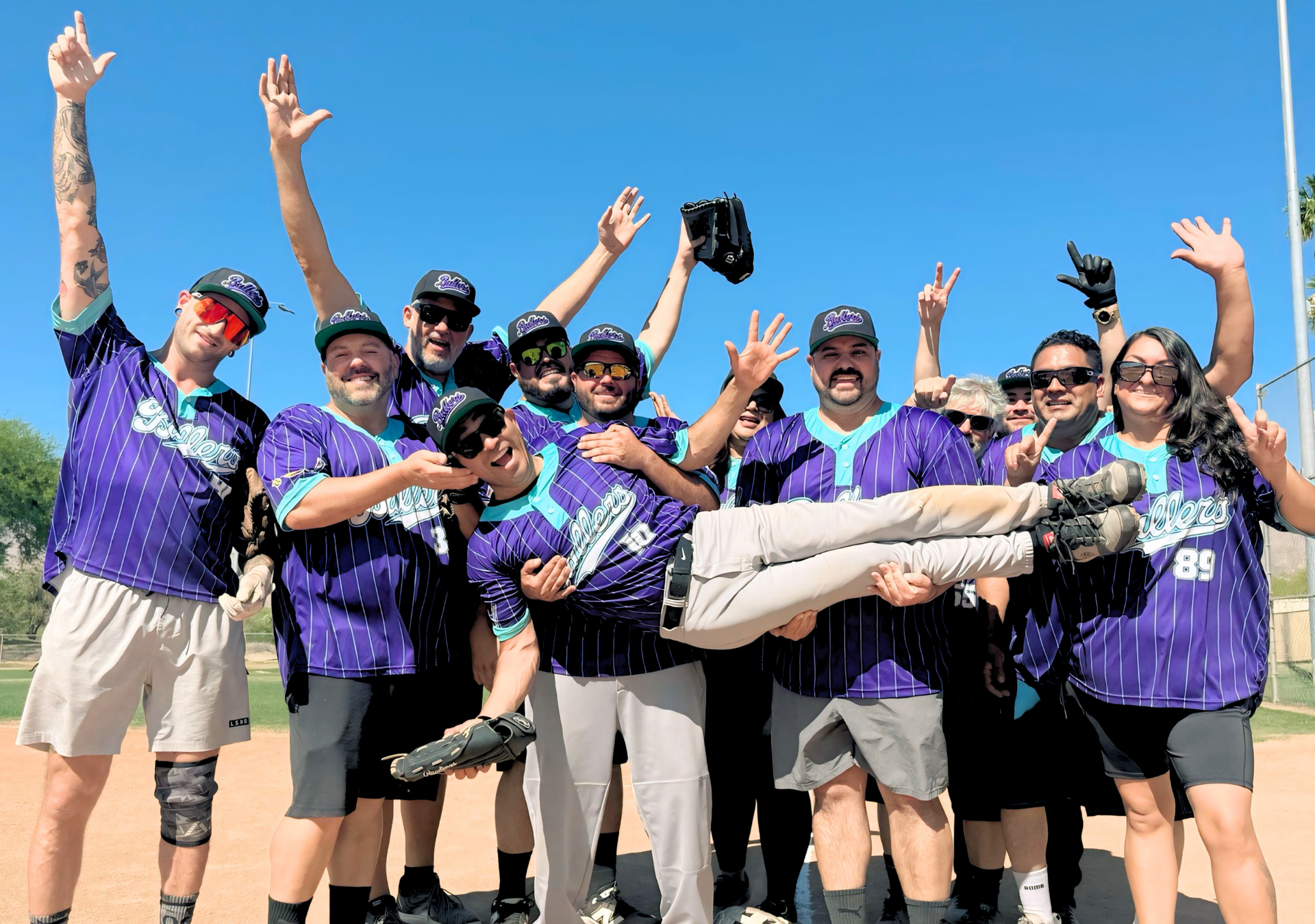 Group of smiling people in purple shirts celebrating outdoors, with one person held horizontally in front.
