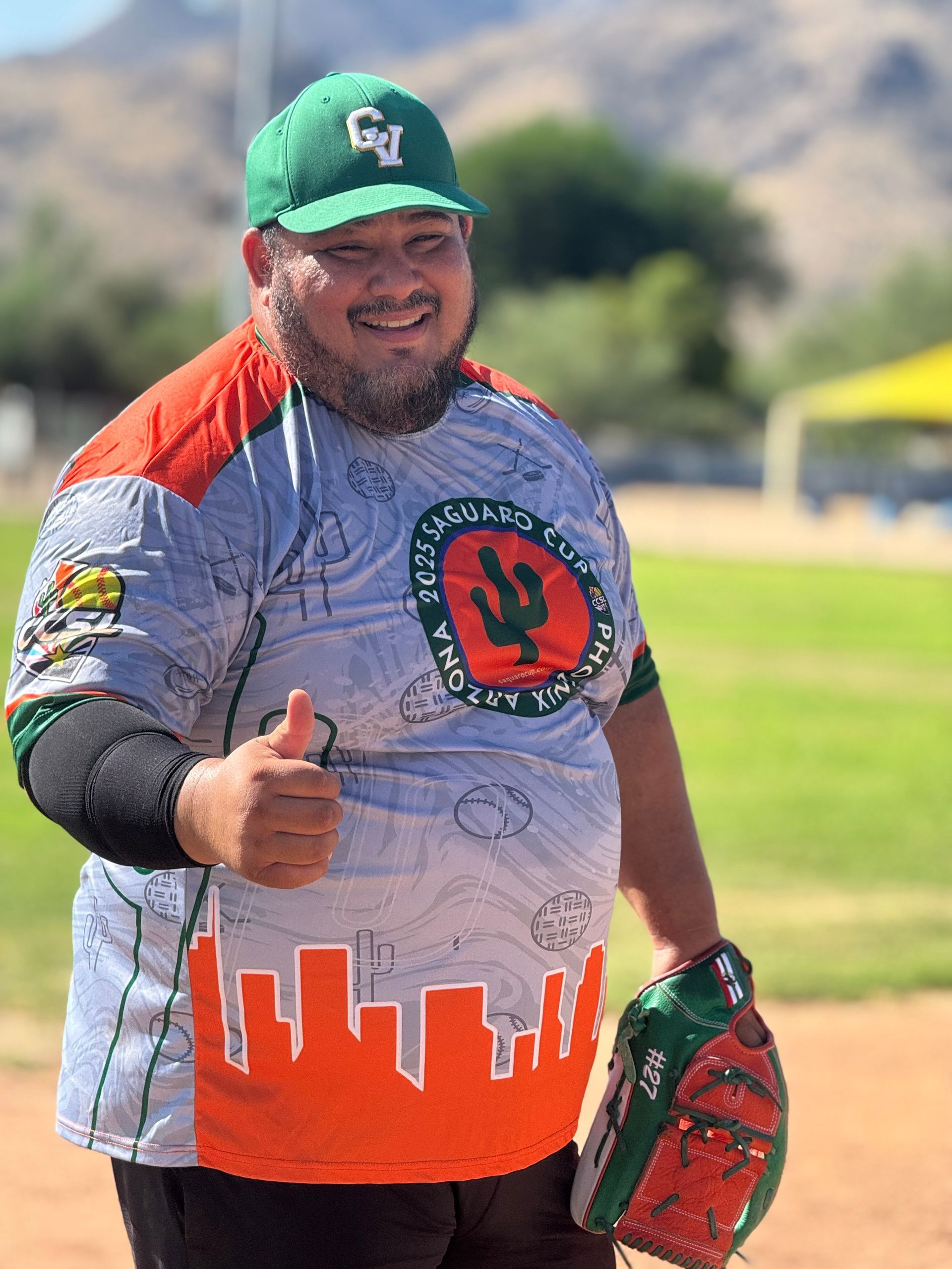 Smiling baseball player in green cap and gray-orange jersey giving a thumbs-up on a field