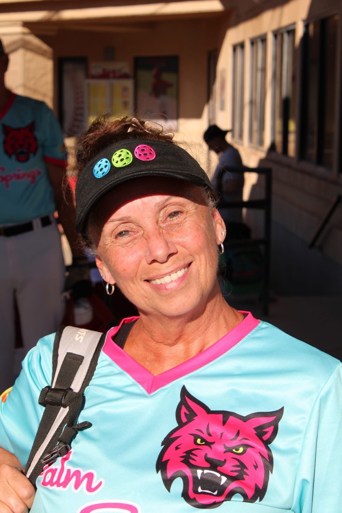 Smiling woman in turquoise jersey and black cap with colorful pins, standing outdoors in sunlight