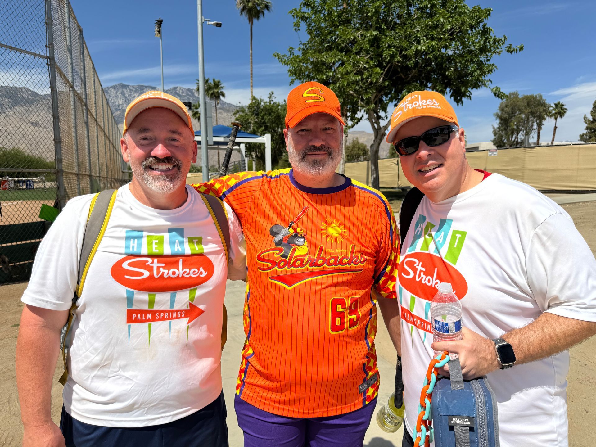 Three people in orange event shirts posing outdoors on a sunny day near a fence and palm trees.