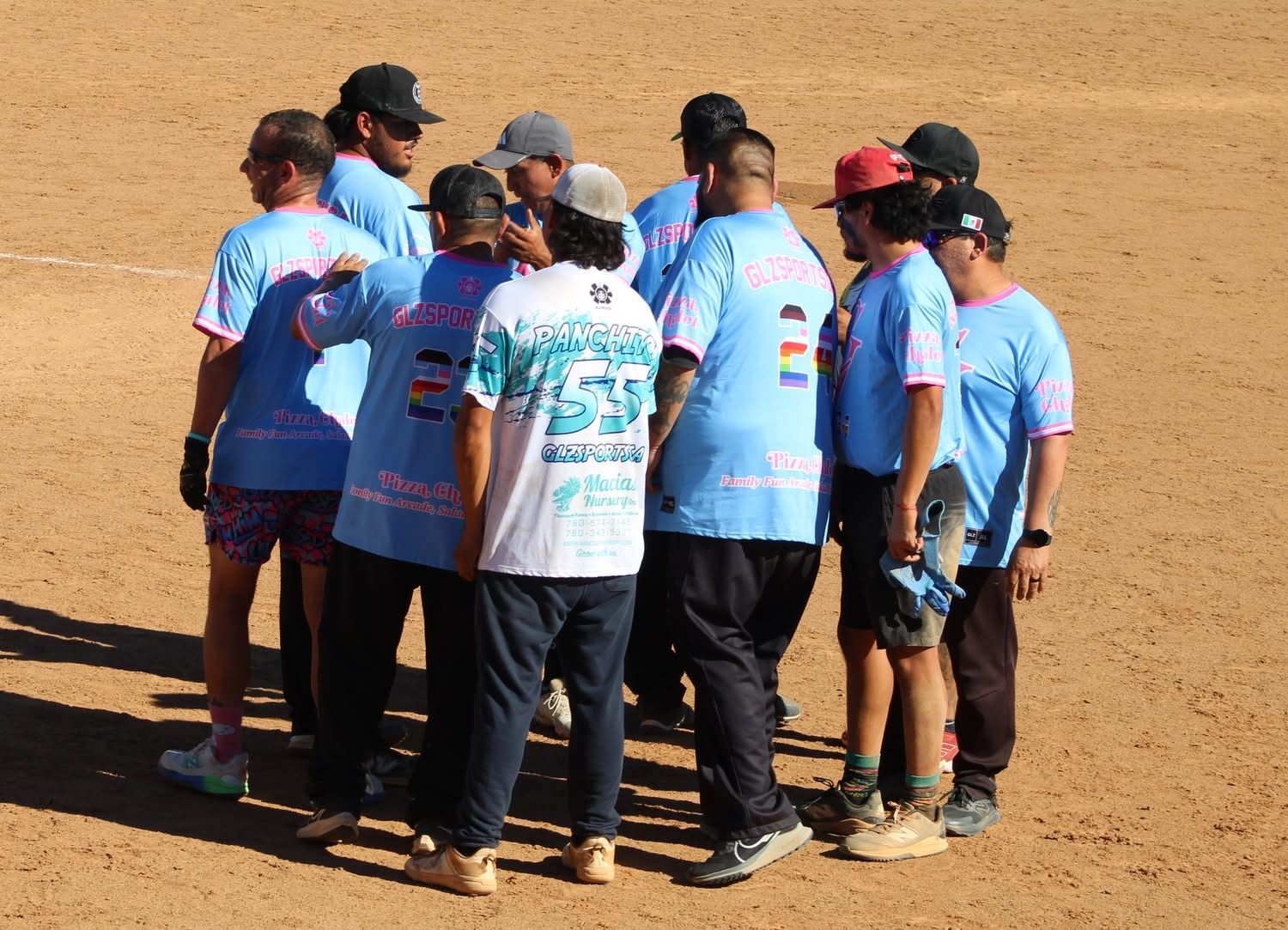 Group of people in light blue shirts huddling together on a sandy outdoor field.