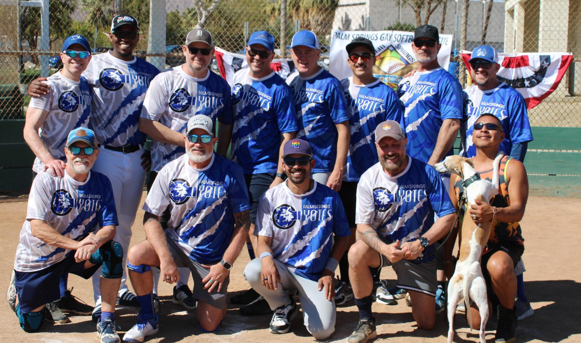 Baseball team in blue jerseys posing outdoors with a trophy and patriotic bunting in the background