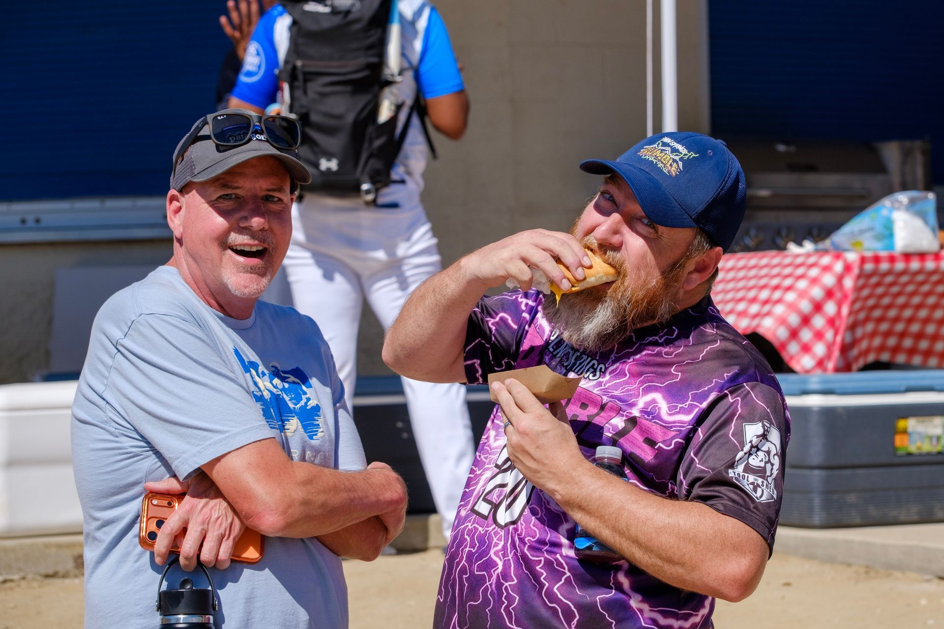Two fans laughing outside a stadium, one in a purple jersey eating food, the other in a gray shirt and cap.