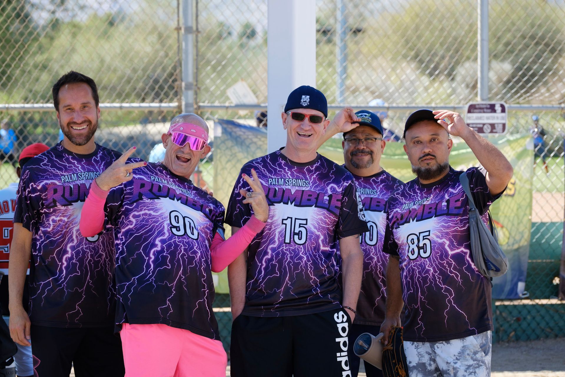 Five softball players in purple jerseys pose on a field, smiling and holding up their hands.