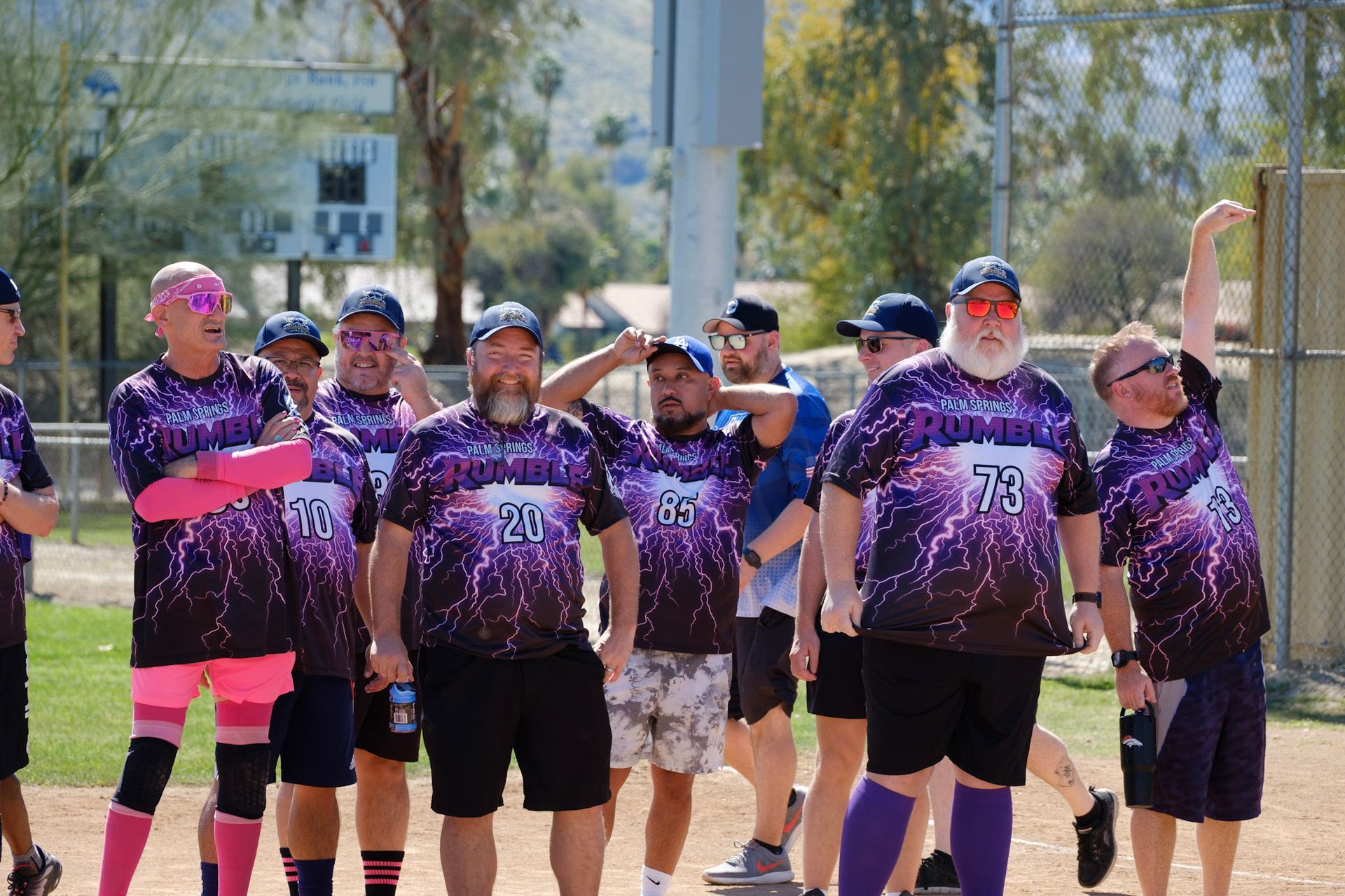 A softball team in purple jerseys stands together outdoors on a sunny field.
