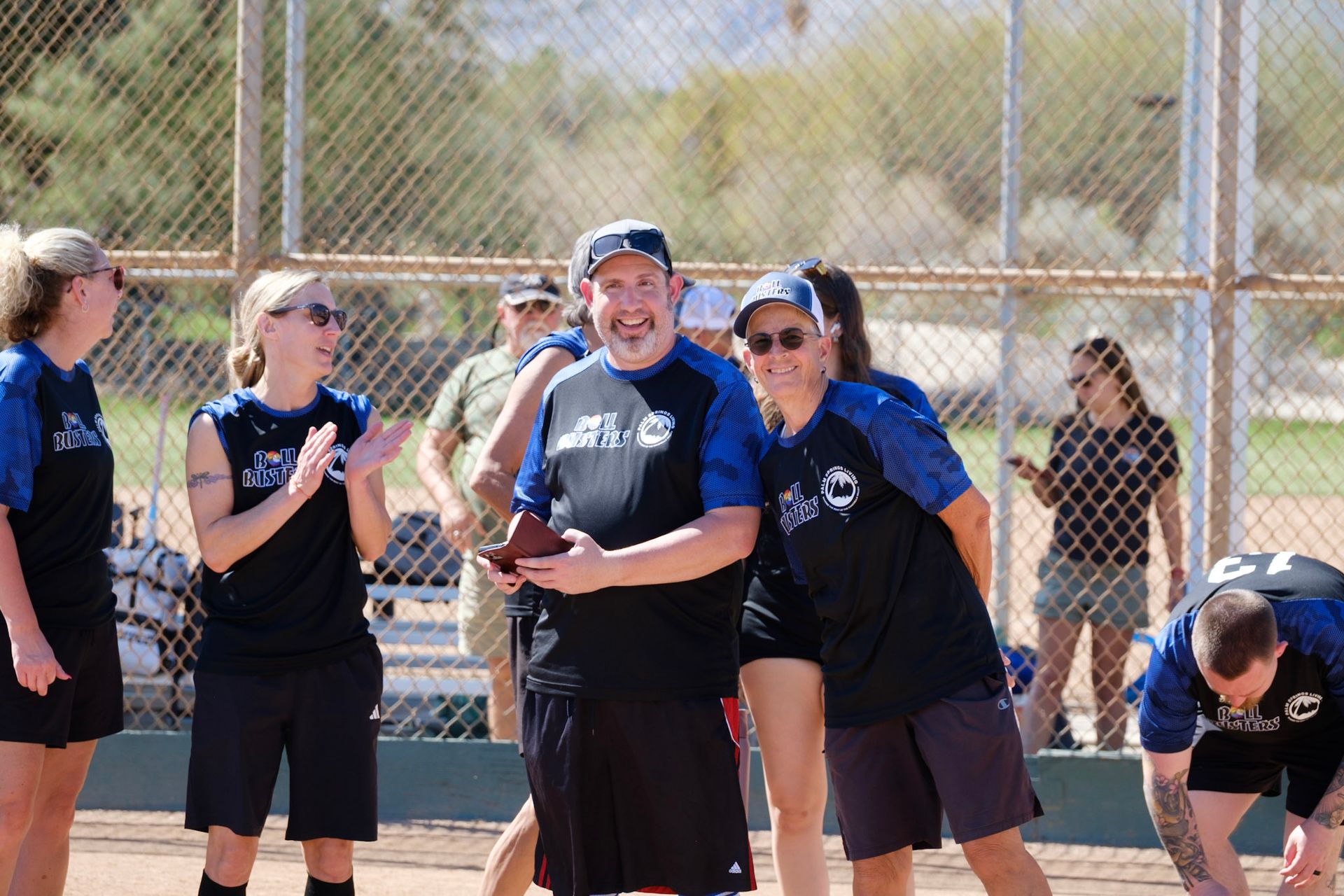 People in black shirts smiling on a softball field behind a chain-link fence