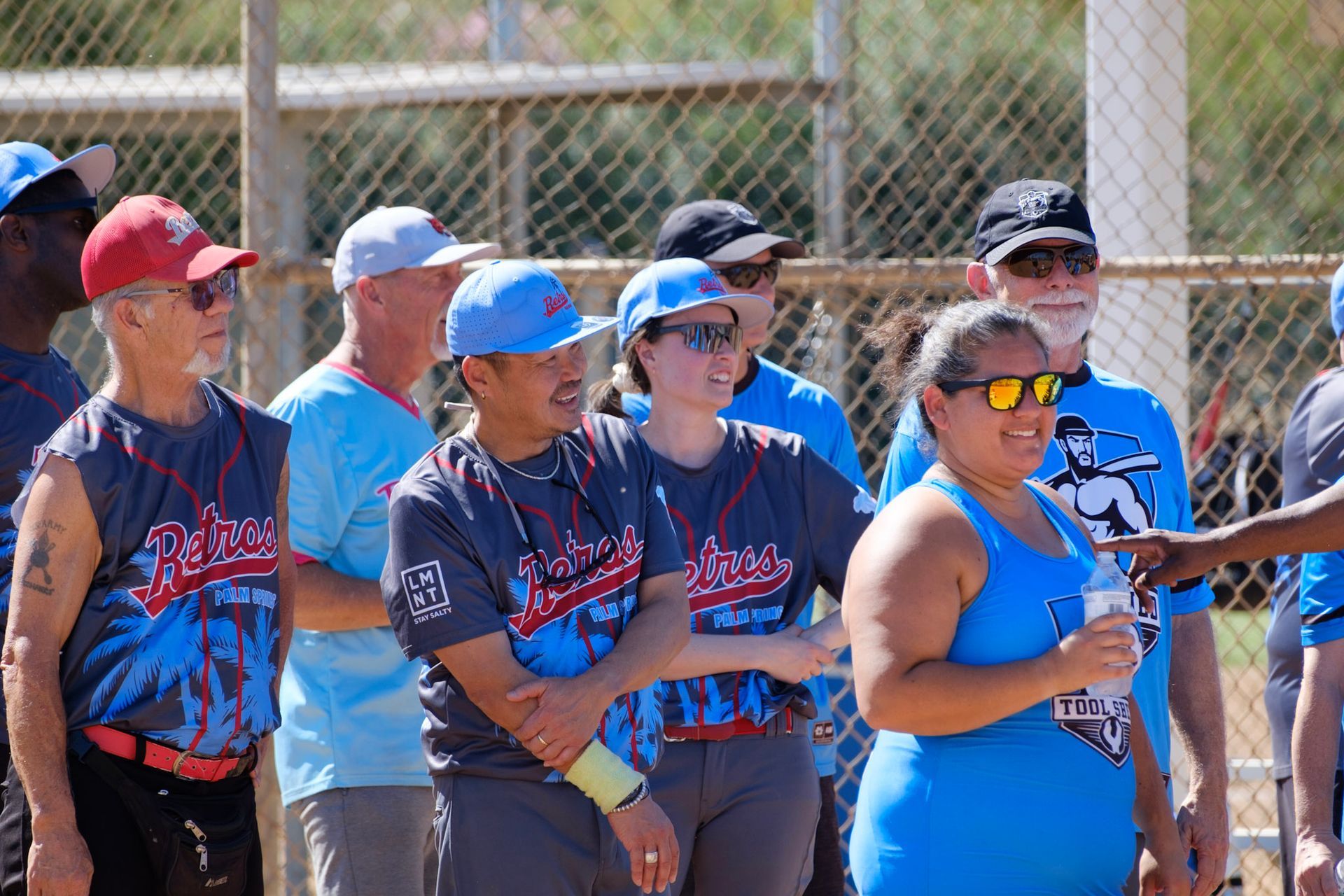 Group of people in matching blue shirts and caps standing outdoors near a fence, some wearing sunglasses.