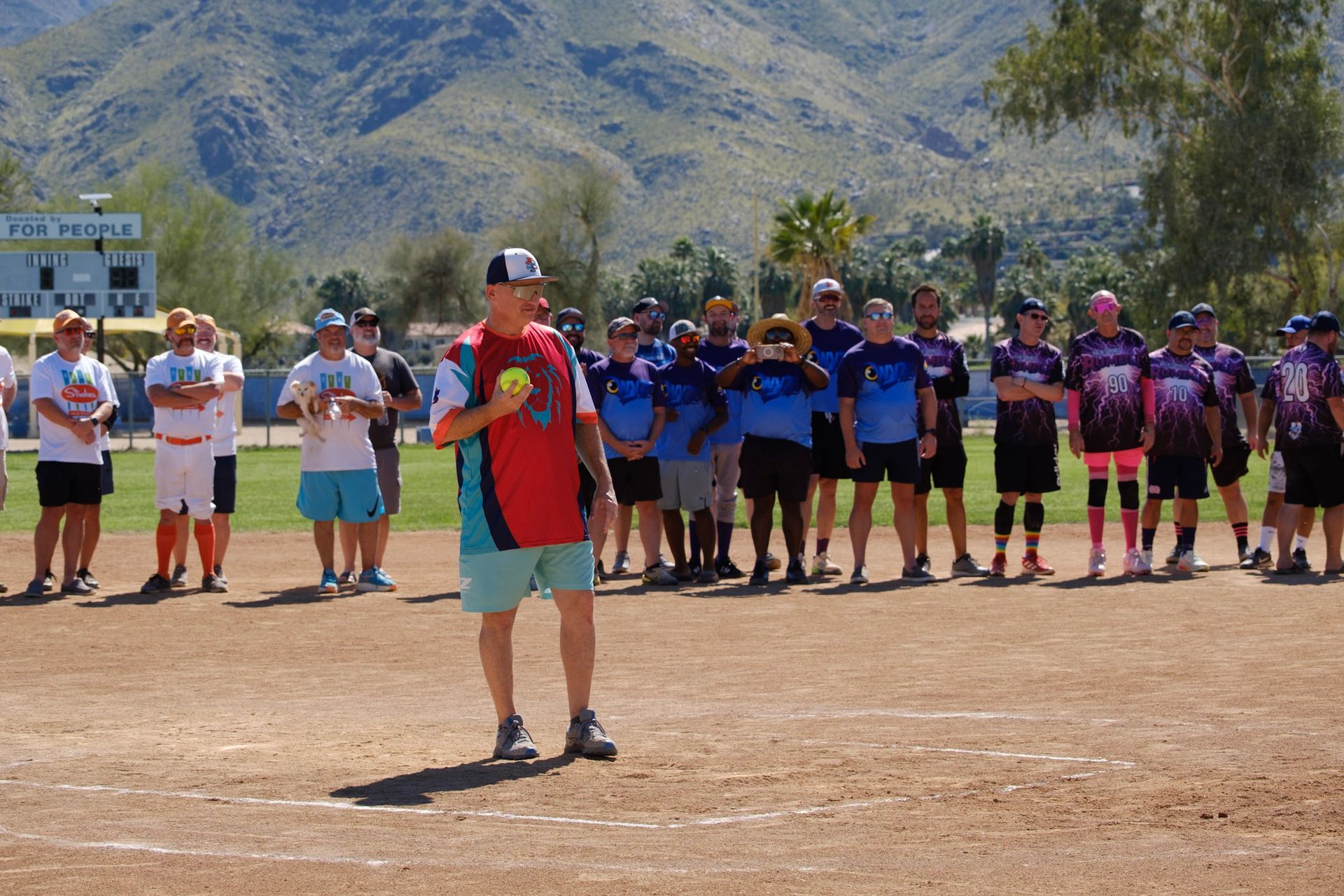 People lined up on a baseball field with mountains in the background; a man in red stands in front holding a microphone.