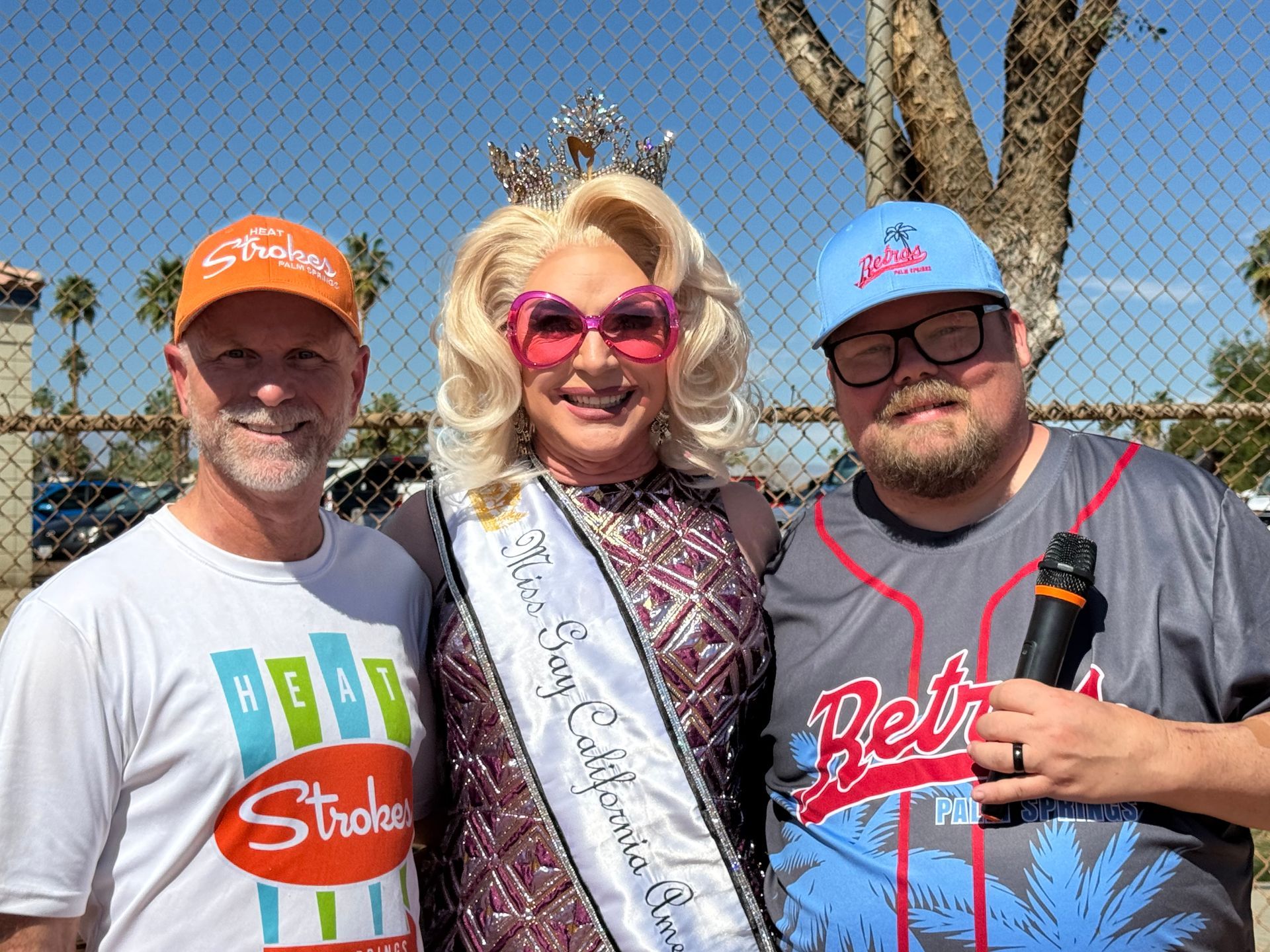 Three smiling people posing outdoors in front of a fence and trees, two wearing baseball caps and one in sunglasses