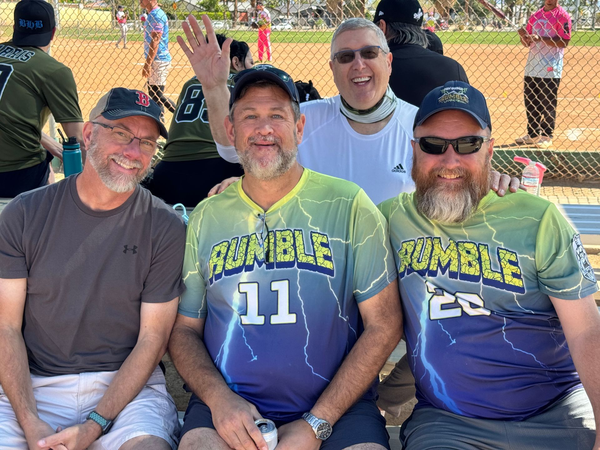 Four men in matching tie-dye jerseys and caps pose smiling on a softball field behind a fence.